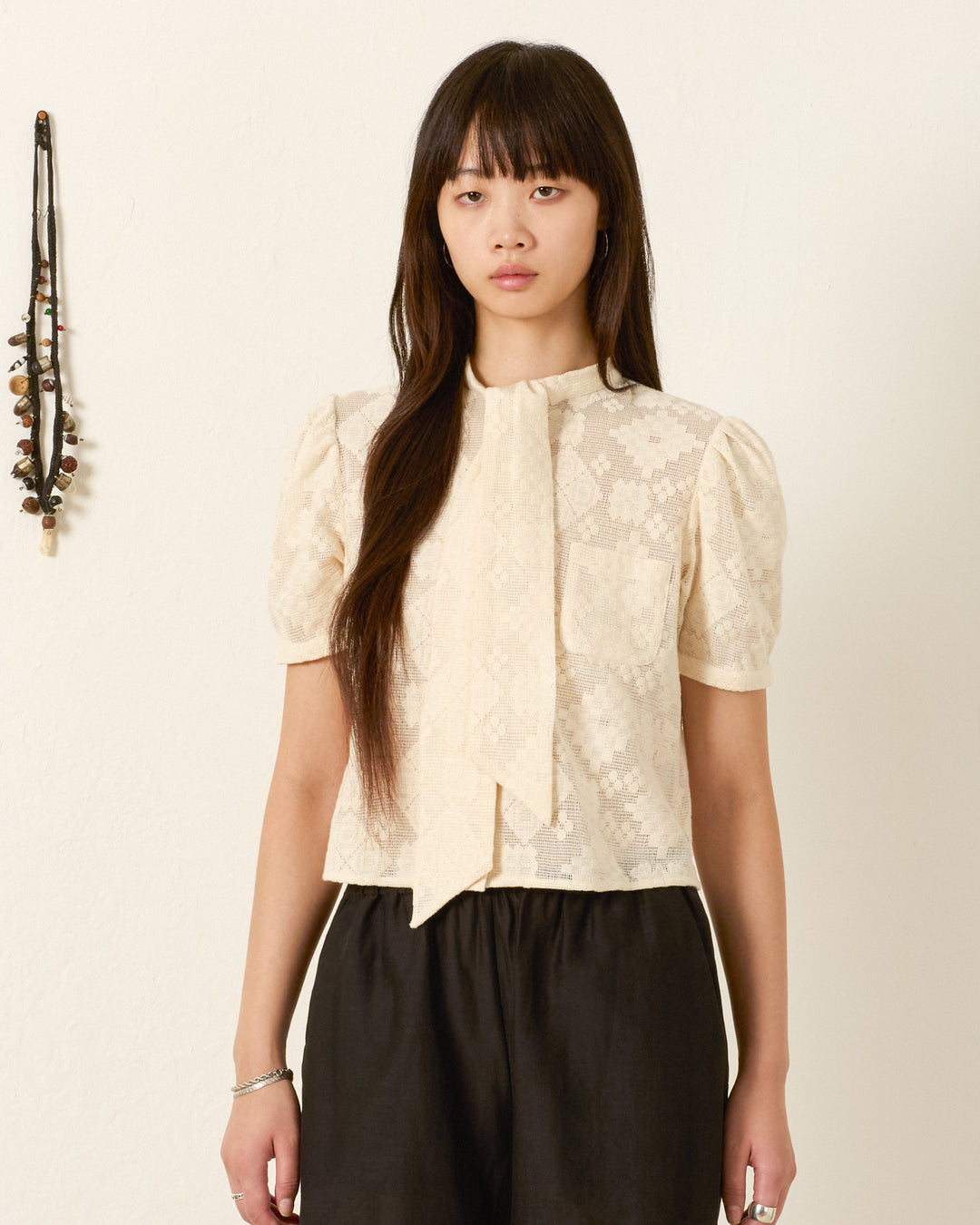 A woman stands against a plain wall, wearing the Chance Ecru Tetris Lace Top with a cropped silhouette and tie detail, paired with black pants. A string of beads hangs on the wall beside her.