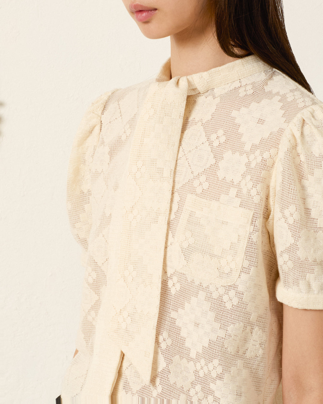 A woman models the Chance Ecru Tetris Lace Top, a light ecru cotton blouse with short sleeves, cropped fit, front tie detail, chest pocket, and lace design, shown from shoulders to waist.