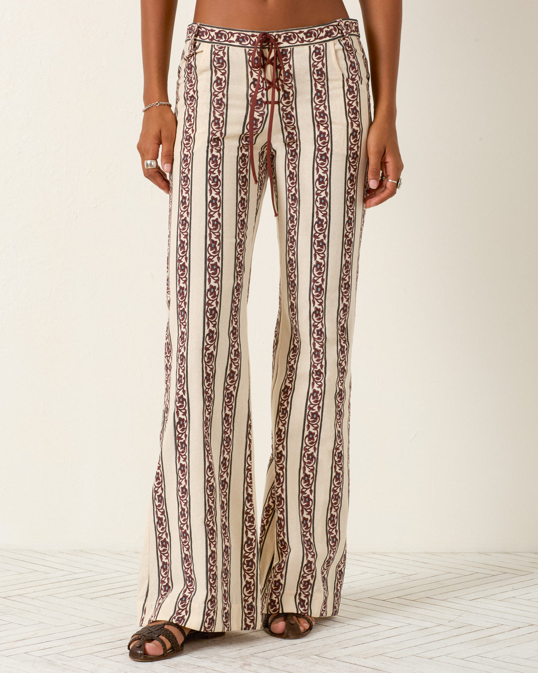 A woman wears the Cooper Climbing Vine Pant—high-waisted, flared khadi cotton pants with vertical maroon and cream geometric stripes, hand-block-printed—standing against a plain light background.