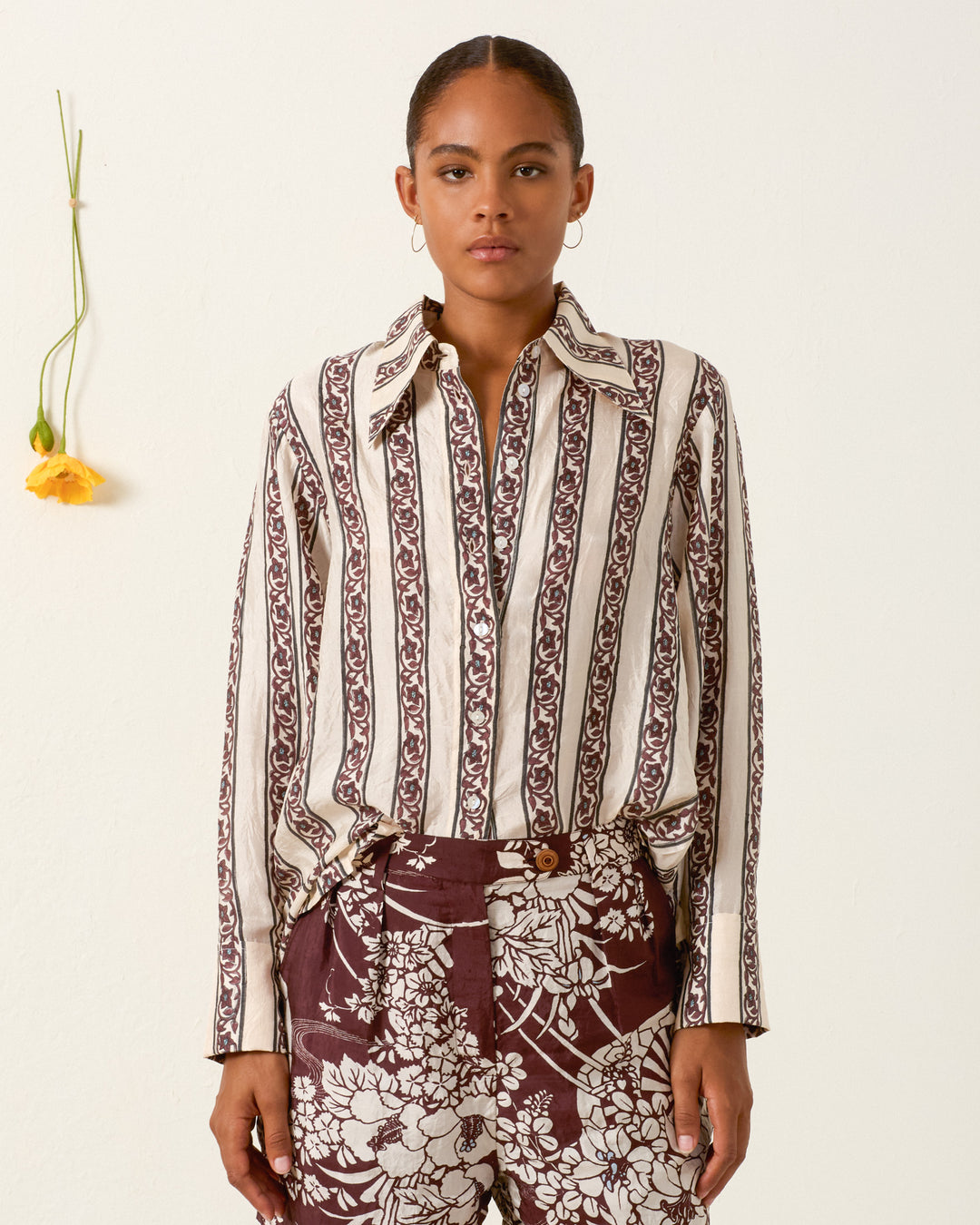 A woman stands against a plain wall, wearing the Dana Climbing Vine Silk Shirt; a single yellow flower hangs on the wall beside her.
