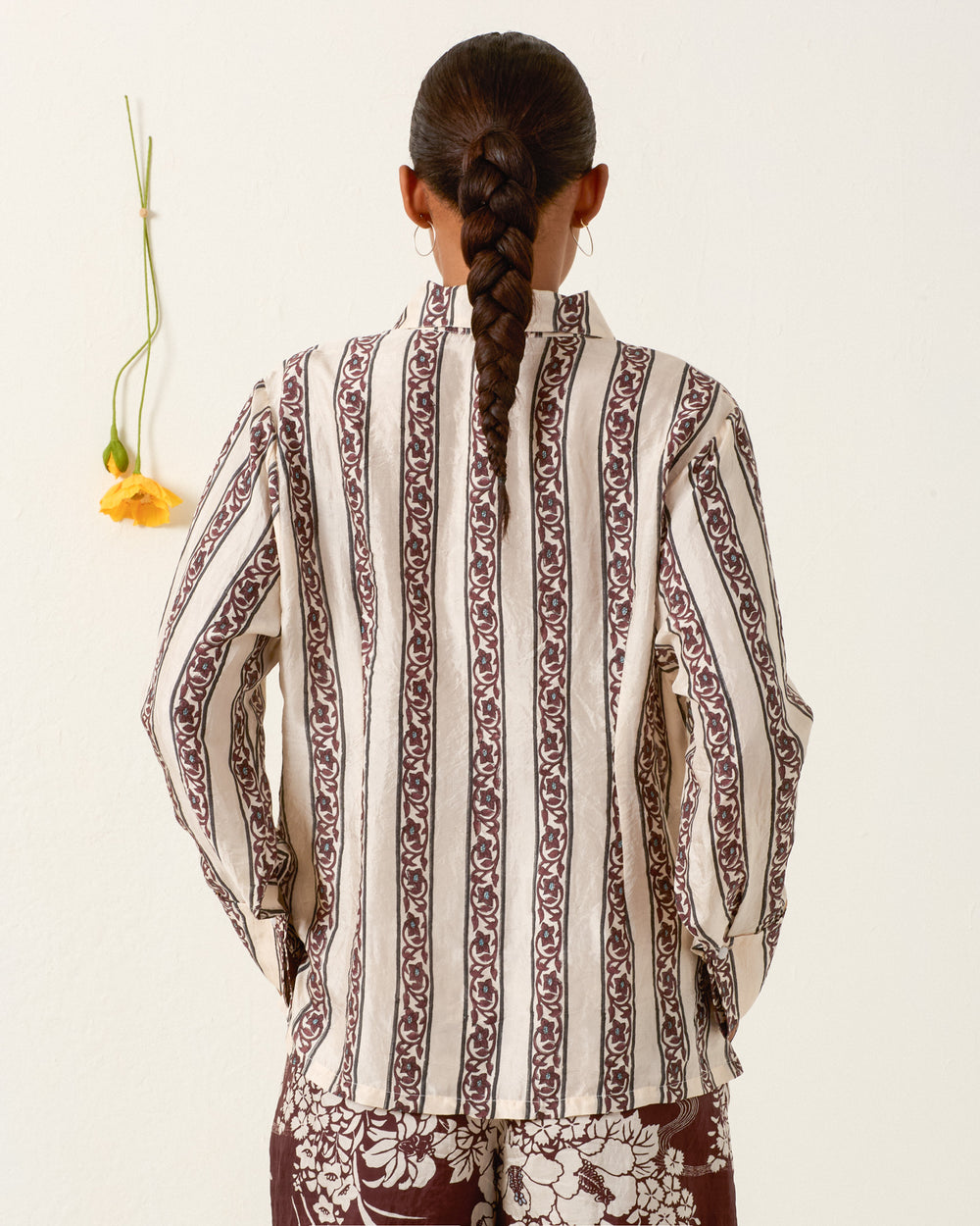 A person with a braided ponytail faces away, wearing the Dana Climbing Vine Silk Shirt with long sleeves and hand-loomed silk, paired with floral pants. To the left, a single yellow flower hangs on the wall.