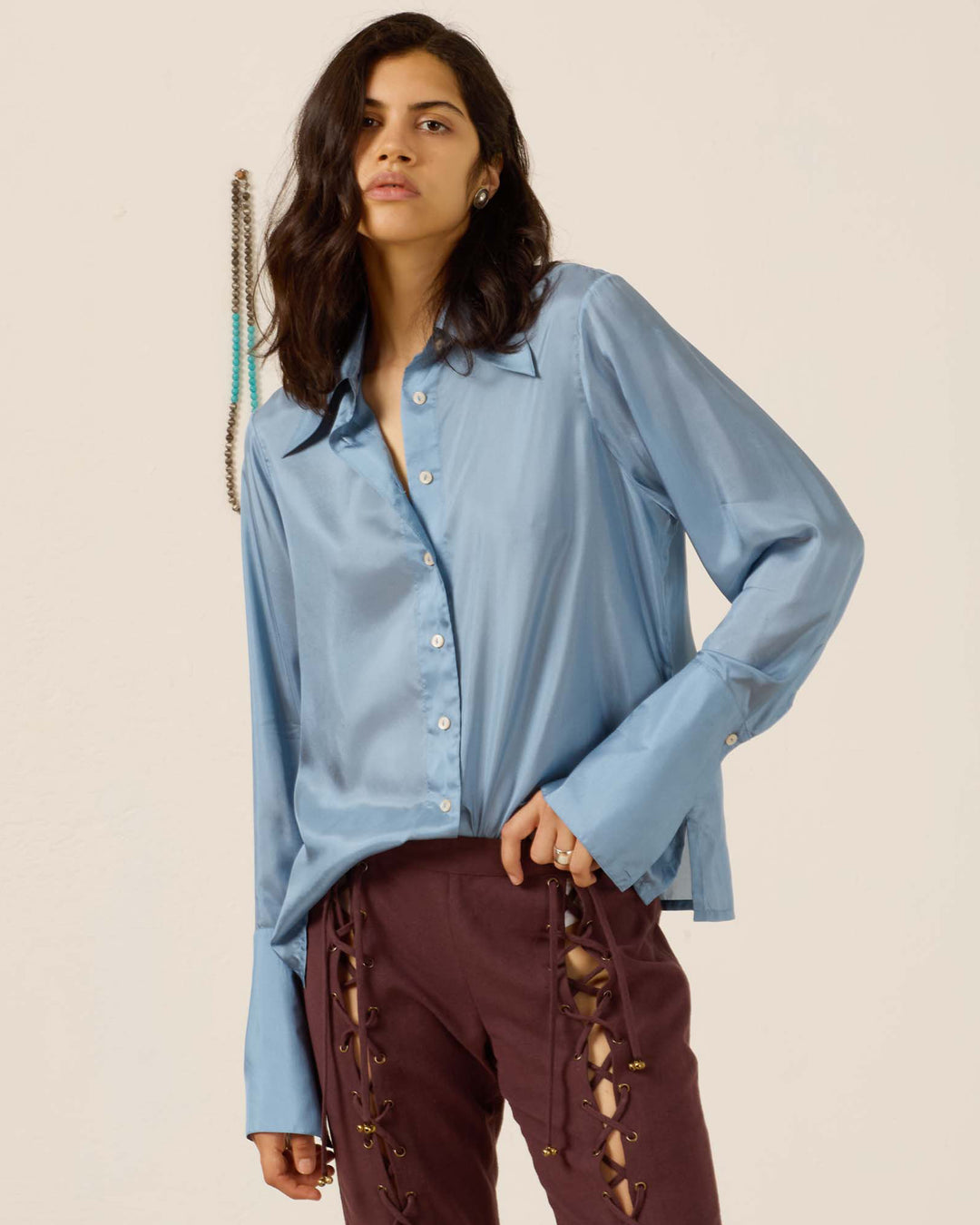 Wearing the Dana Chambray Silk Shirt—a loose, light blue, vintage-inspired top with a ’70s dagger collar—and burgundy lace-up pants, a person stands against a plain background.
