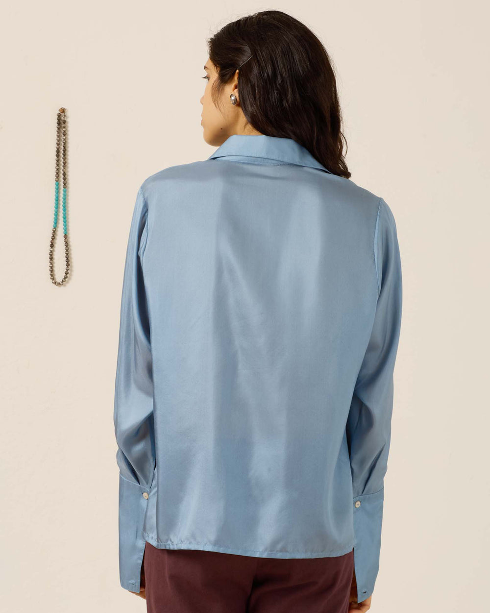 A person with long dark hair, seen from behind, wears the Dana Chambray Silk Shirt—light blue, hand-loomed silk with a ’70s dagger collar—and maroon pants. A beaded necklace hangs on the wall to the left.