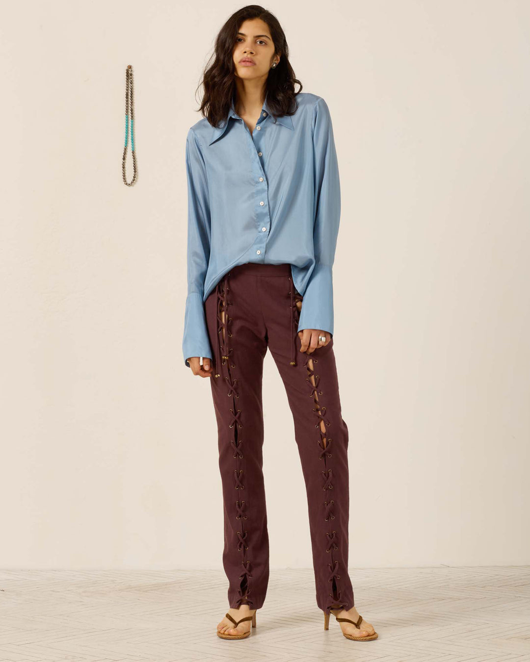A woman stands indoors wearing the Dana Chambray Silk Shirt, a vintage-inspired light blue button-up with a subtle ’70s dagger collar, paired with maroon lace-up pants and heeled sandals. A beaded necklace hangs on the wall nearby.