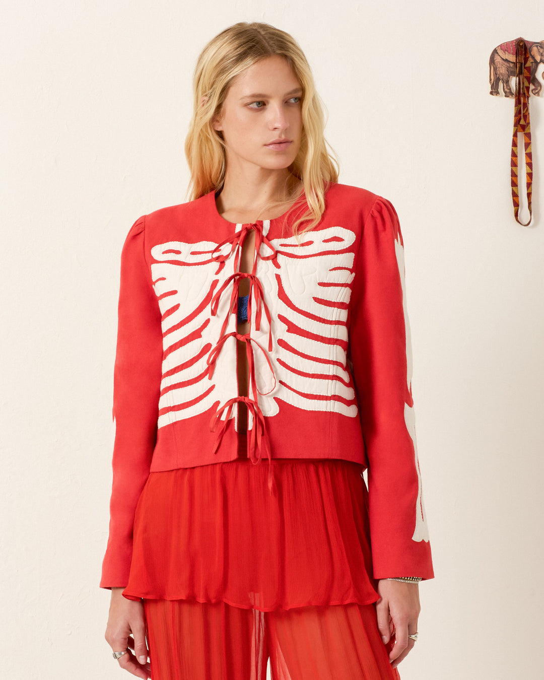 A woman wears the Dorian Ibis Bone Jacket, a red cropped silhouette featuring a white skeleton motif on the front, while looking to the side against a plain background.