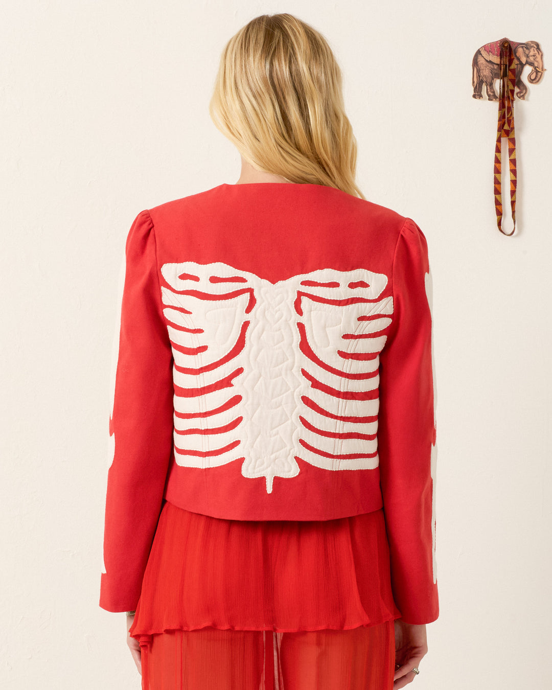 A person with long blond hair wears the Dorian Ibis Bone Jacket—a red, textured cotton cropped jacket with a white skeleton motif on the back—standing in front of a white wall with a decorative elephant hanging.