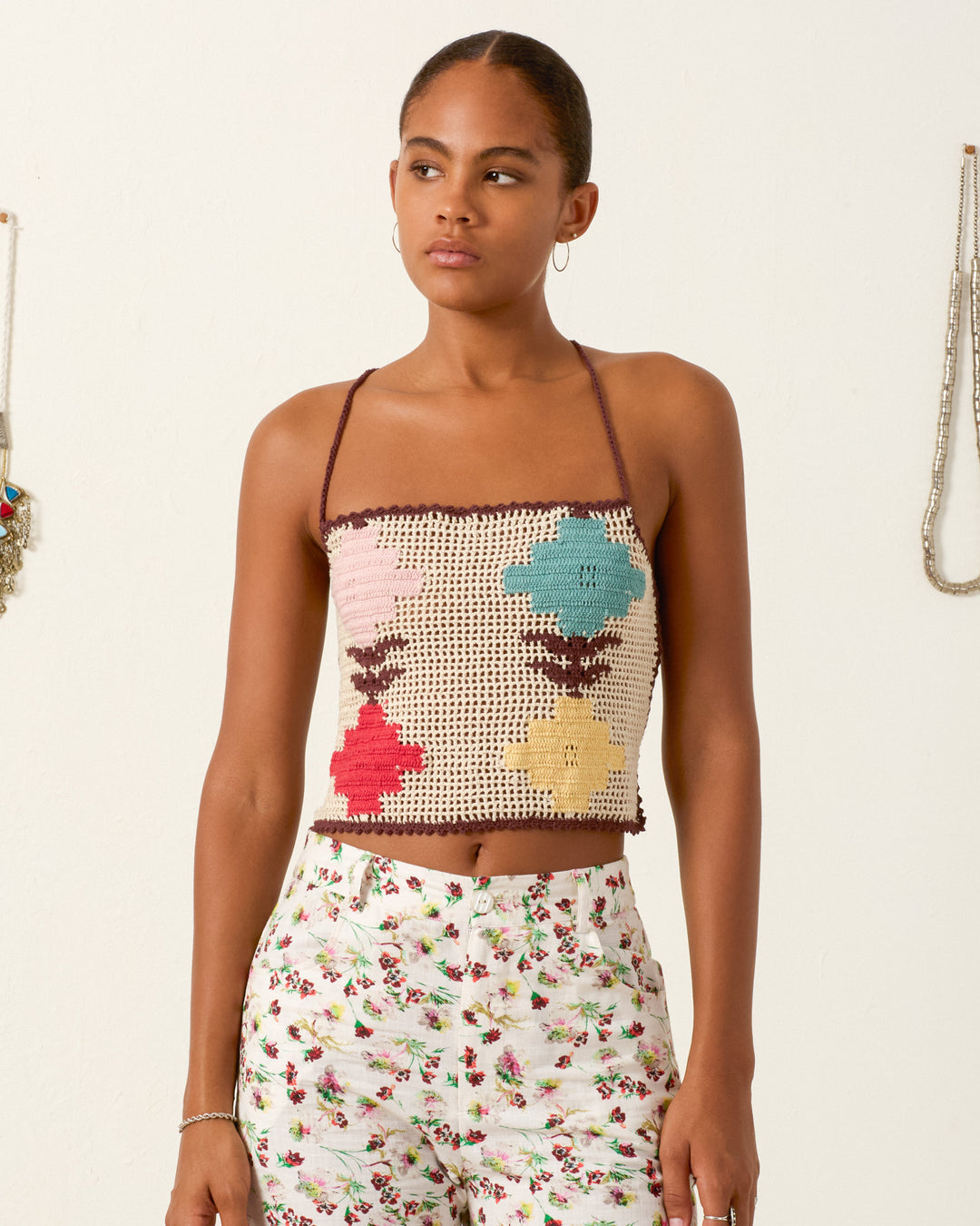 A woman wears the Eliza Daisy Chain Top, a hand-crocheted cotton piece with colorful flower patterns, paired with white floral pants and standing against a plain wall with hanging necklaces in the background.