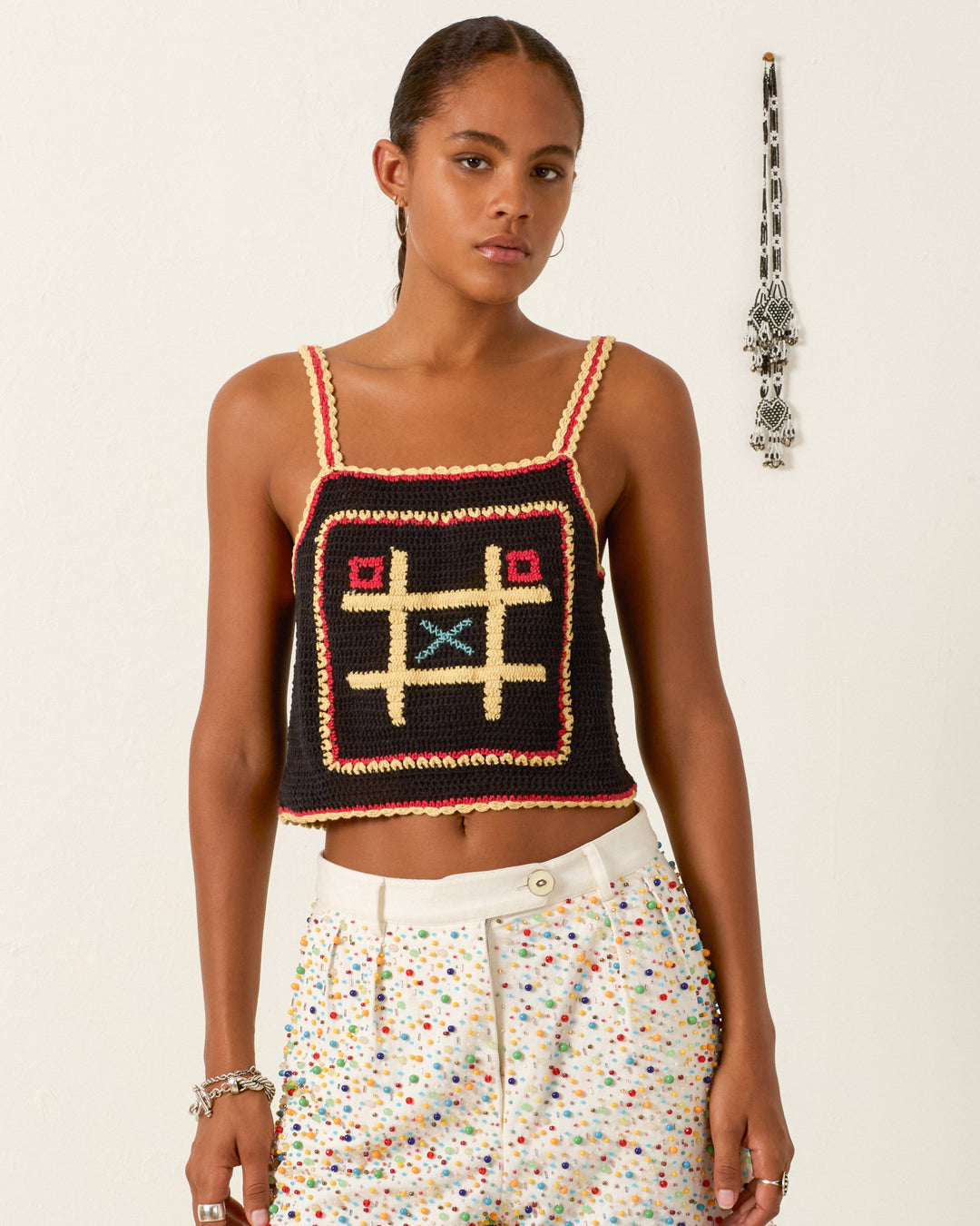 A woman wears the Elide Tic Tac Toe Top, an artisan-made hand-crocheted cotton piece with a cropped silhouette, paired with colorful dotted white pants as she stands against a white wall.