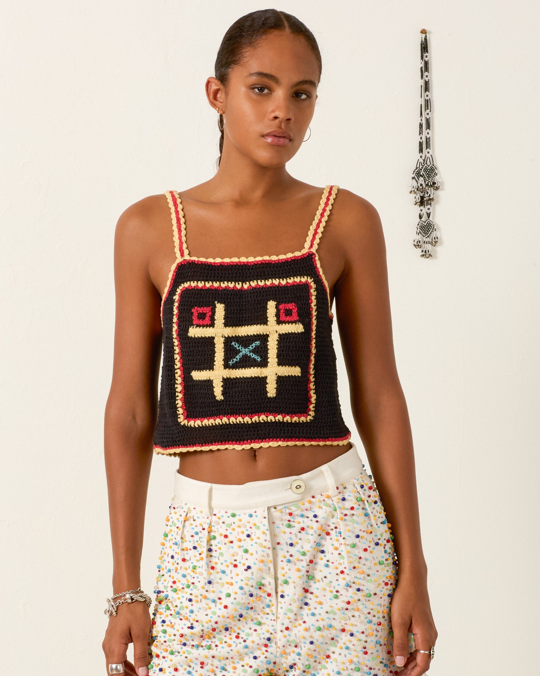 A woman wears the Elide Tic Tac Toe Top, an artisan-made hand-crocheted cotton piece with a cropped silhouette, paired with colorful dotted white pants as she stands against a white wall.