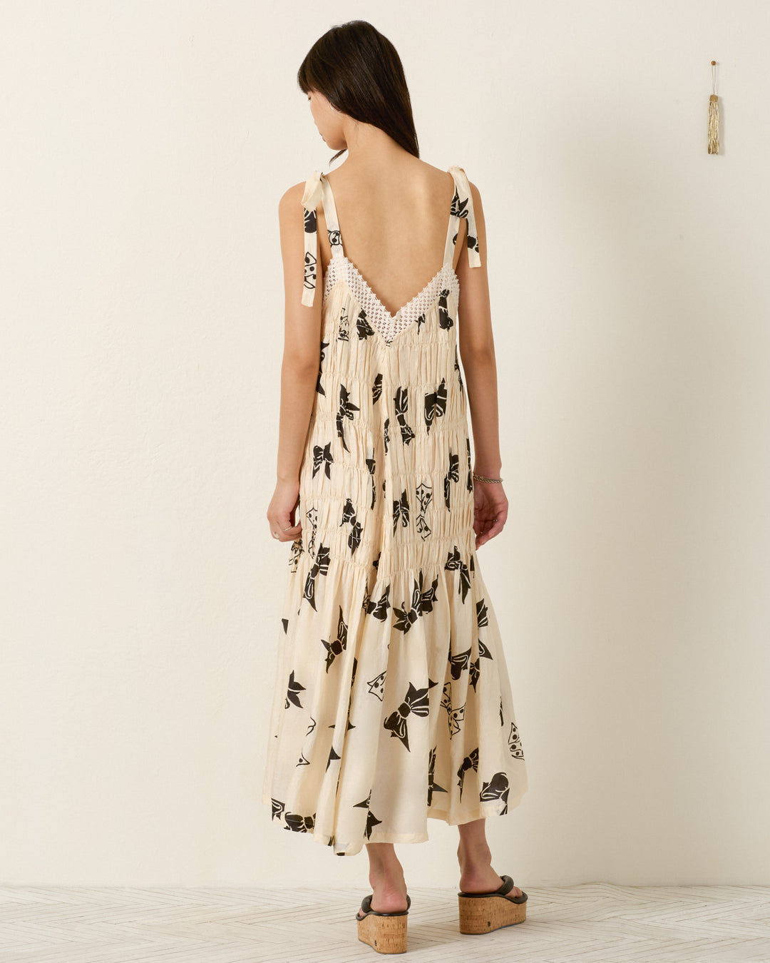 A woman faces away, wearing the Ellina Black Tie Silk Dress in a cream shade with black floral prints and tie straps, paired with platform sandals.