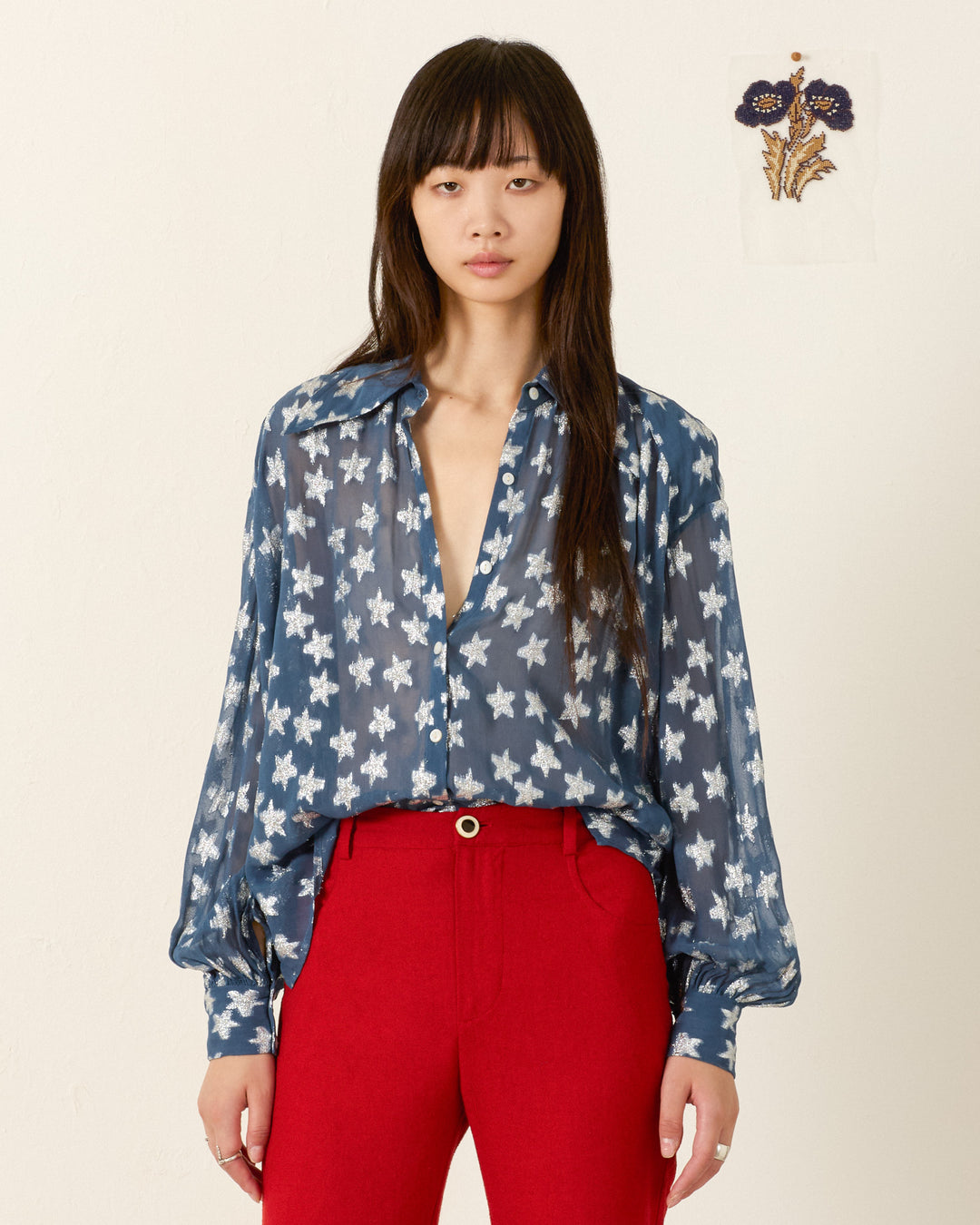 A woman stands against a plain wall, wearing the Emery Starlite Shirt—a semi-sheer blue blouse with white star patterns—and red trousers. A small floral artwork hangs on the wall behind her.