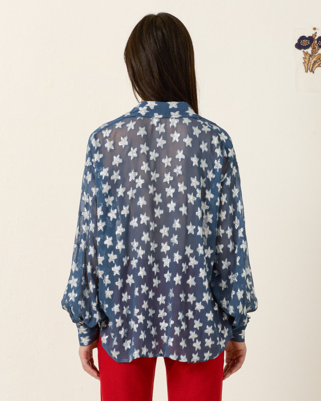 A person with long dark hair faces away, wearing the Emery Starlite Shirt—a metallic silver fil-coupé blouse with white stars—and red pants, standing against a plain light wall.
