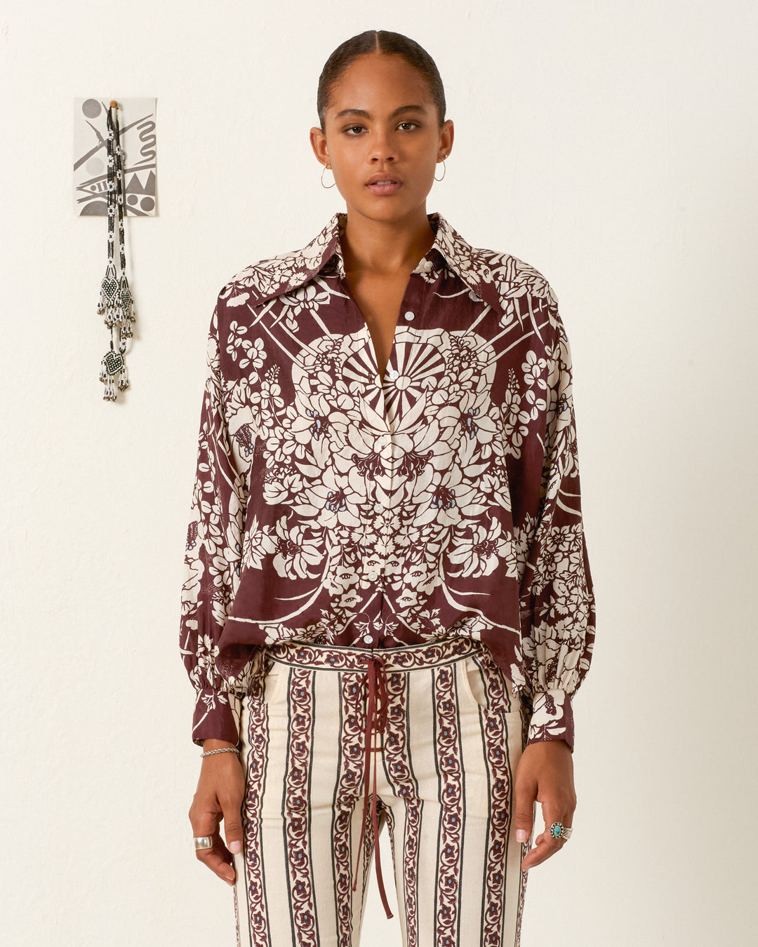 A woman stands against a plain wall wearing the Emery Auburn Spoke Silk Shirt in maroon and white stripes. A decorative wall hanging is visible behind her.