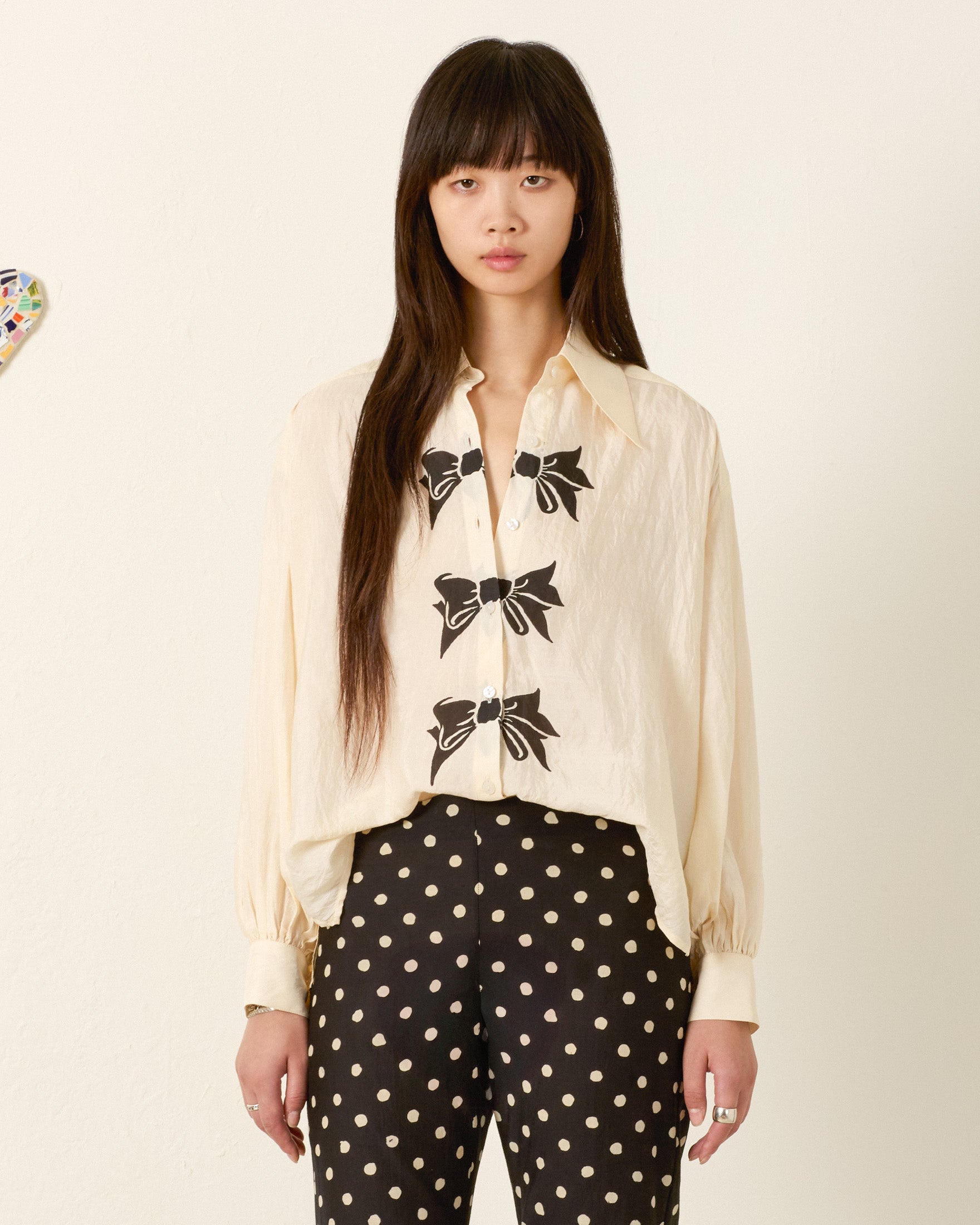 A woman stands against a plain wall wearing the Emery Black Tie Silk Shirt paired with black pants featuring white polka dots.