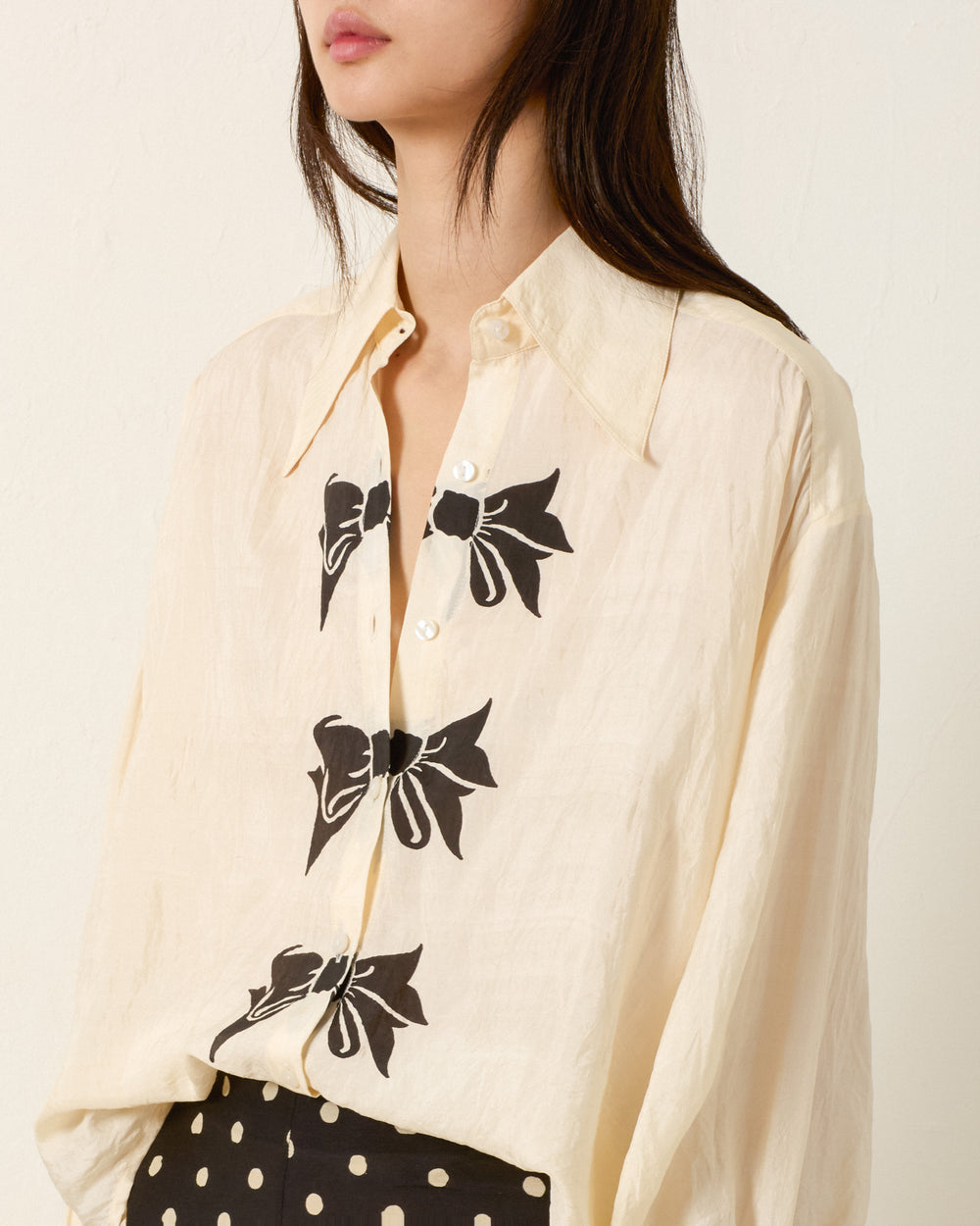 A woman wears the Emery Black Tie Silk Shirt, an artisan block-printed cream button-up with three trompe l’oeil bows on the front, paired with black polka dot pants.