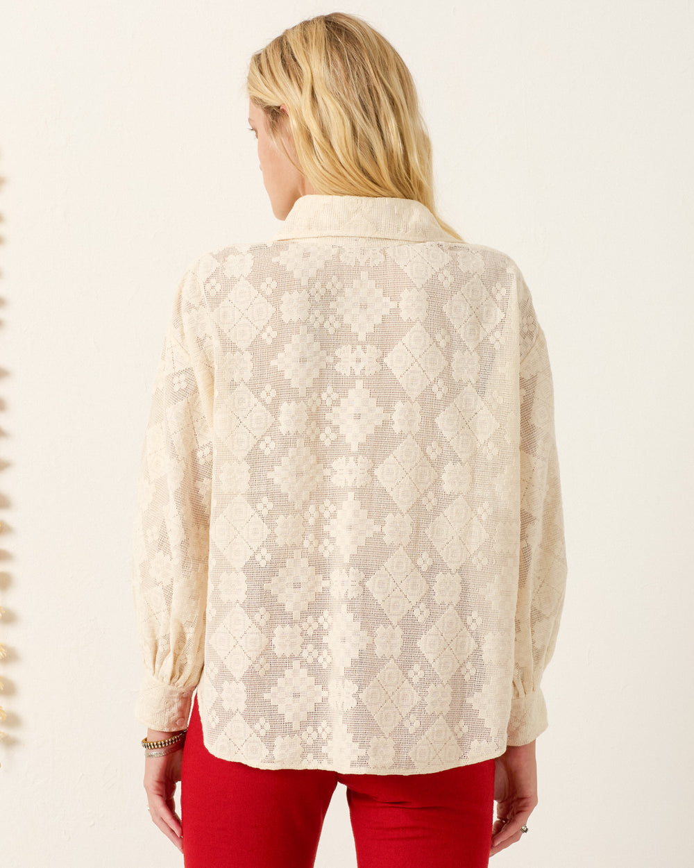 A person with long blonde hair, wearing the Emery Ecru Tetris Lace Shirt and red pants, stands with their back to the camera against a plain light background.