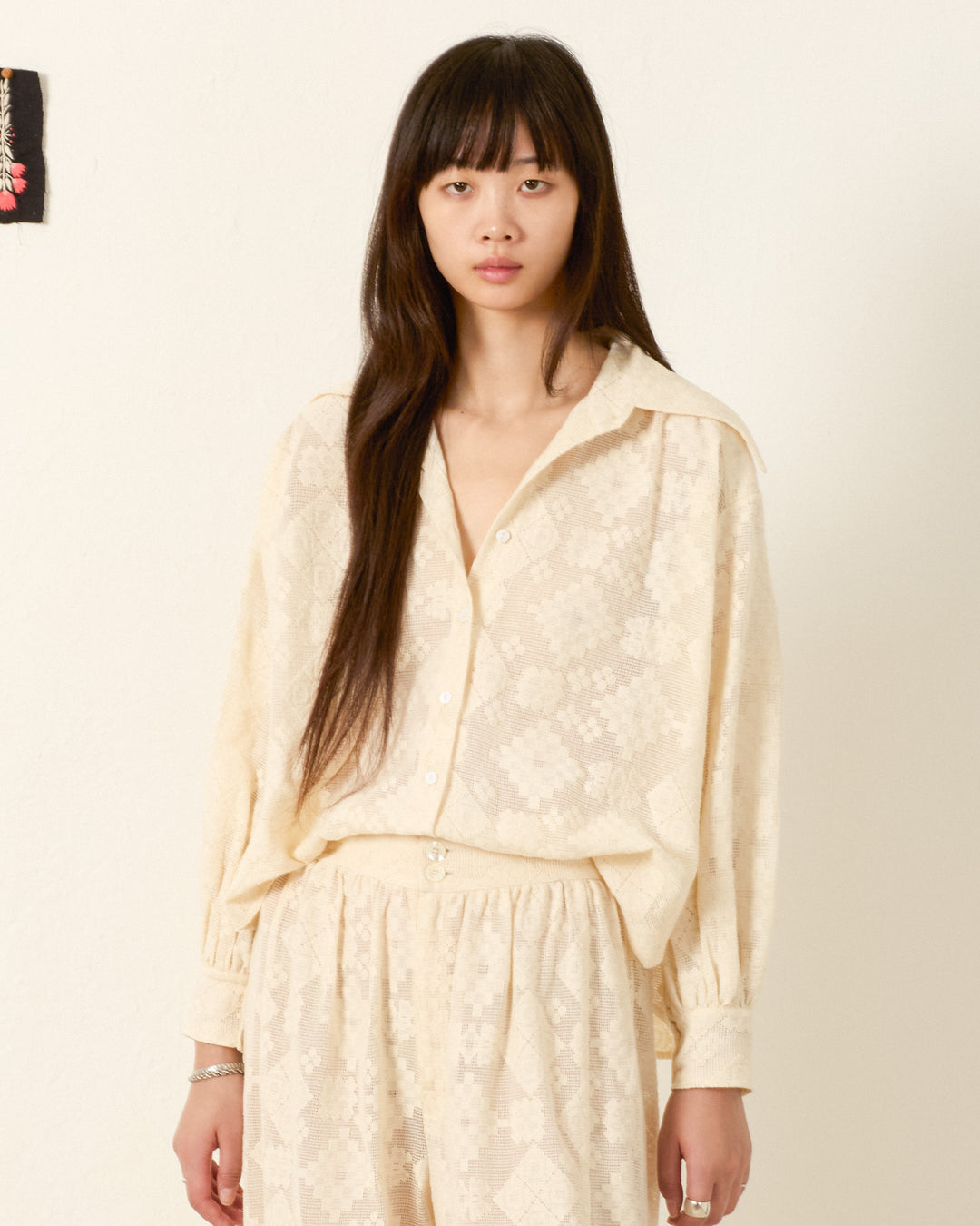 A woman with long dark hair wears the Emery Ecru Tetris Lace Shirt and a matching skirt, standing against a plain light wall.