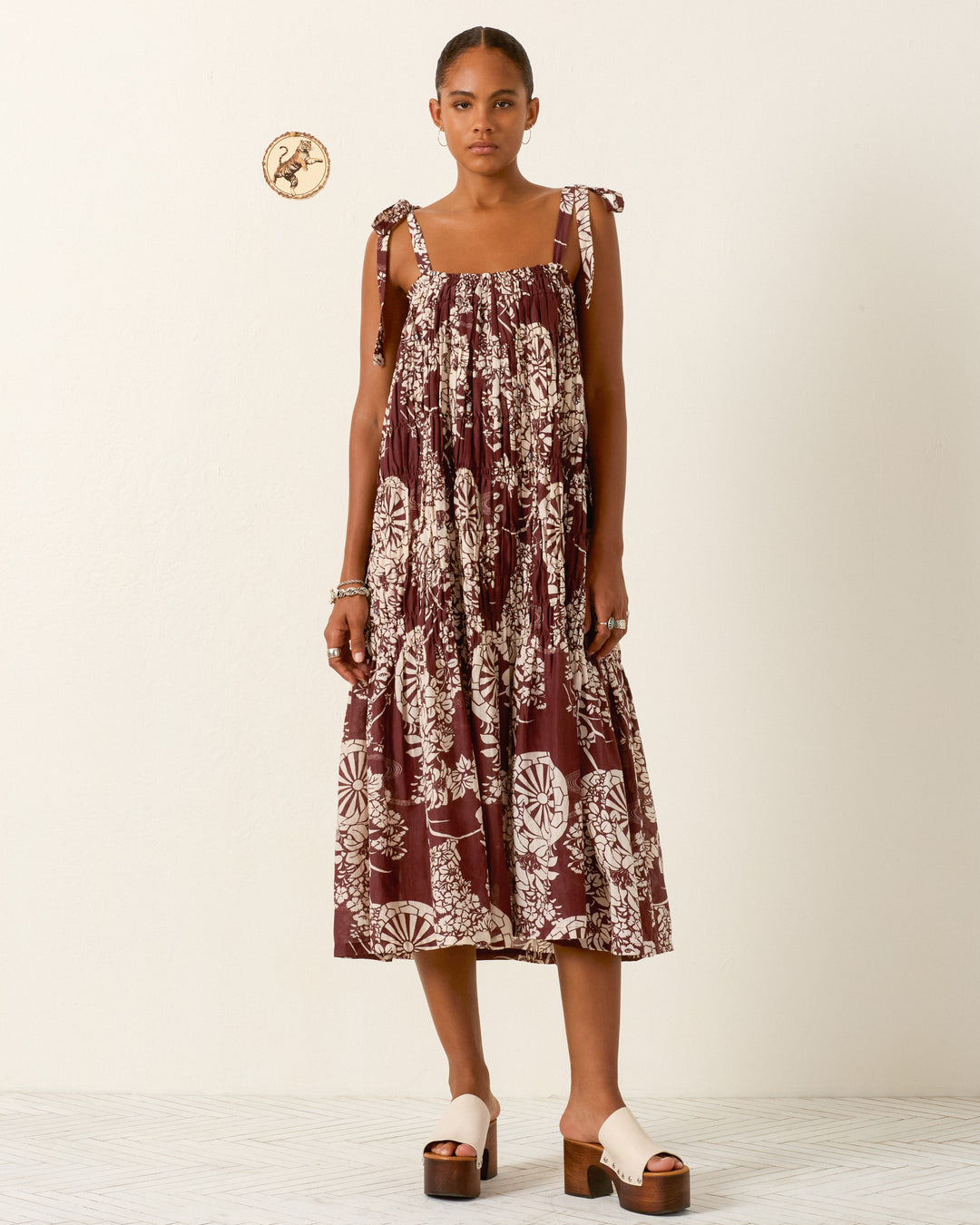 A woman stands against a plain background wearing the Emrys Auburn Spoke Dress—a maroon, floral-patterned cotton-silk maxi with ruched tiers and tie straps—paired with cream platform sandals.