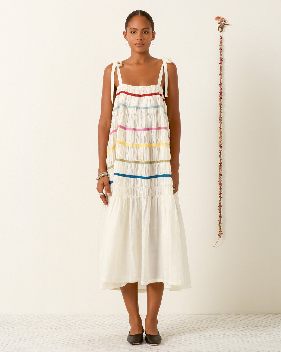 A woman stands indoors wearing the Emrys Ivory Aurora Dress, a sleeveless hand-loomed silk piece with colorful horizontal stripes, paired with black flats and her arms relaxed at her sides.