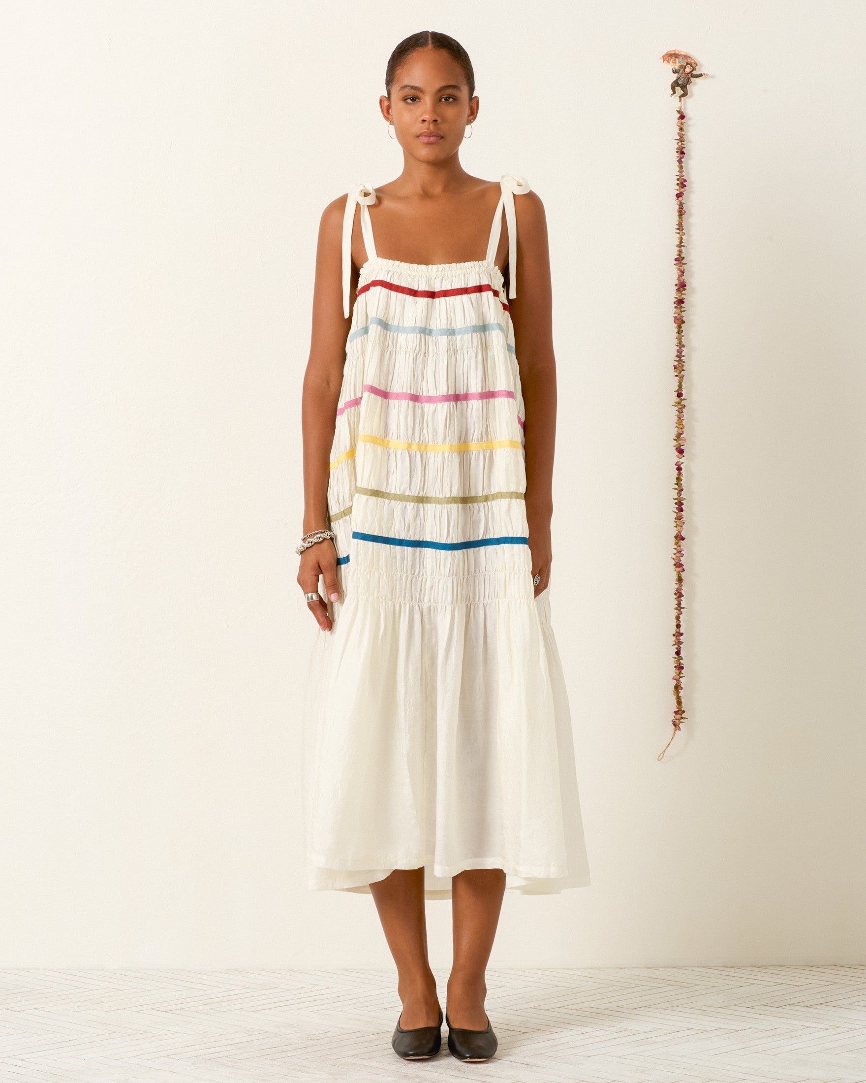 A woman stands indoors wearing the Emrys Ivory Aurora Dress, a sleeveless hand-loomed silk piece with colorful horizontal stripes, paired with black flats and her arms relaxed at her sides.