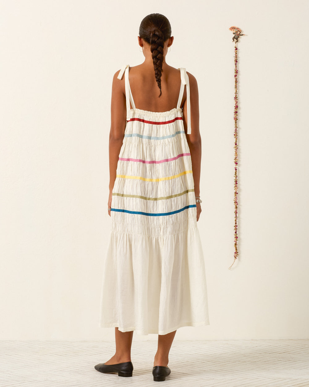 A woman with a braid stands indoors facing away from the camera, wearing the Emrys Ivory Aurora Dress—a sleeveless, white hand-loomed silk maxi dress with colorful horizontal stripes.