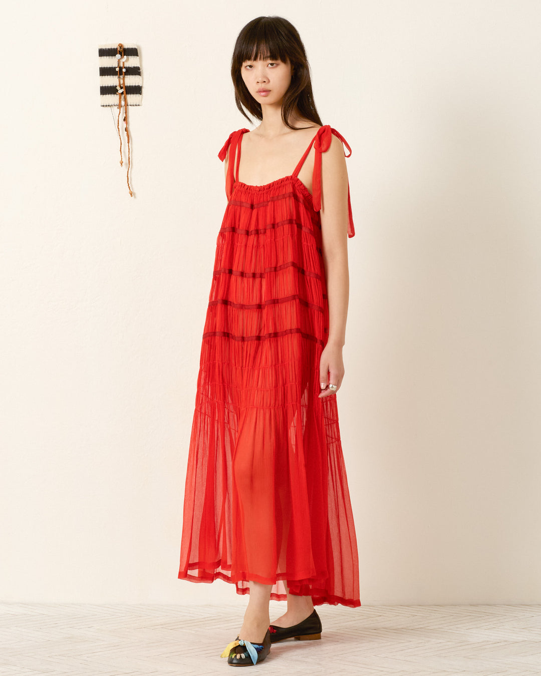 A woman stands indoors against a plain wall, wearing the Emrys Ibis Chiffon Dress—a sheer red maxi with tie shoulder straps and ruched tiers—paired with black flats. A small woven decoration hangs nearby.