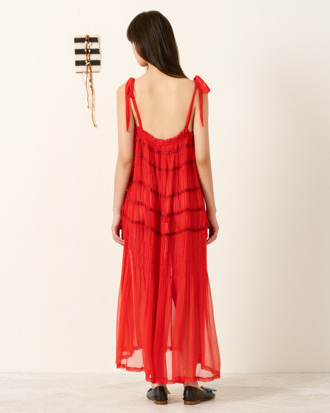 A woman stands indoors on a light floor near a white wall with a woven hanging, wearing the Emrys Ibis Chiffon Dress—red cupro-chiffon with tie shoulder straps and ruched tiers.