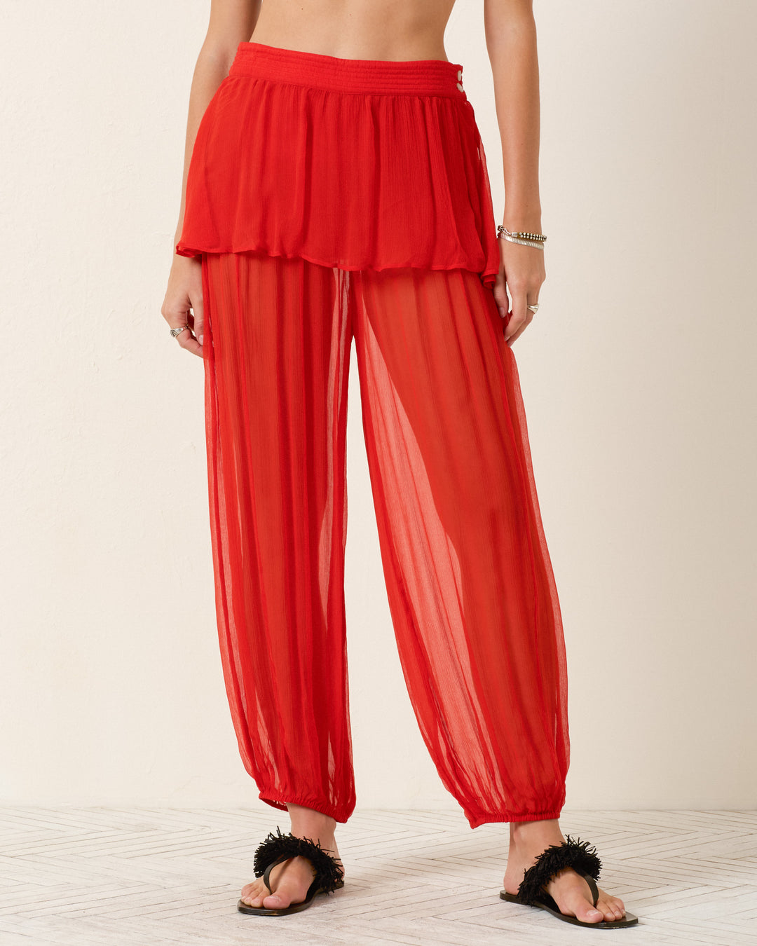 A person wearing the Evangeline Ibis Chiffon Pant in semi-sheer red with a solid skirt overlay, black sandals, and a silver bracelet stands against a plain light background.