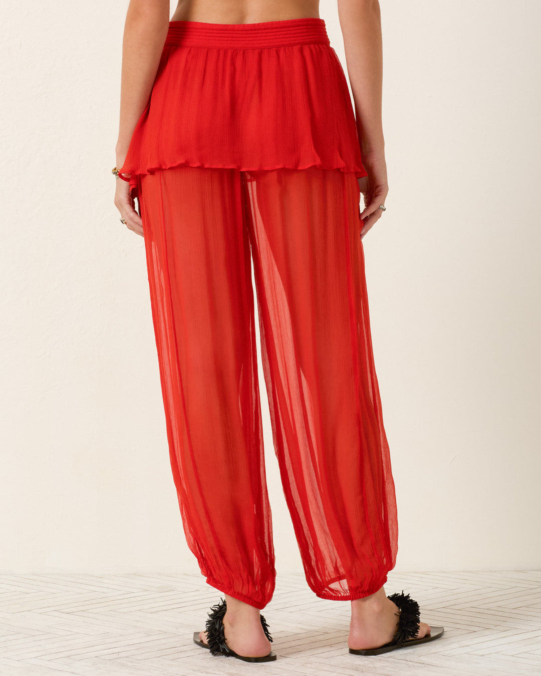 Model shown from the back in Evangeline Ibis Chiffon Pant—semi-sheer red cupro-chiffon with a skirt overlay waistband—styled with black flat sandals against a plain light backdrop.