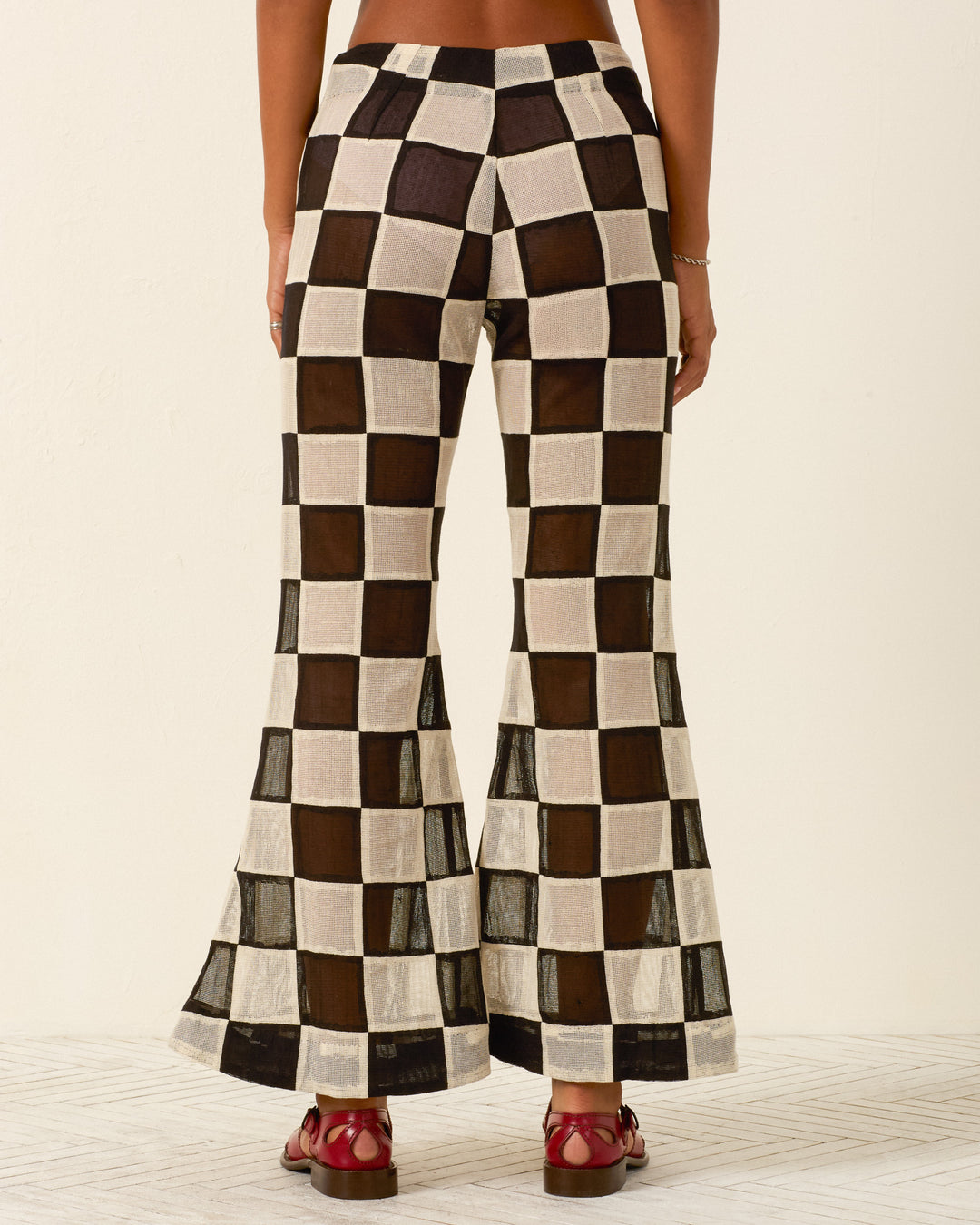 A person wearing the Farah Harlequin Pant, featuring semi-sheer cotton with a black and white checkered patchwork pattern, seen from the back and styled with red sandals.