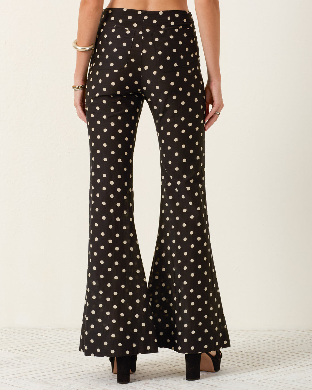 A person is seen from the back wearing the Farah Blanc Speckle Pant, high-waisted with a black and white polka-dot flare design, styled with black platform heels and gold bracelets.