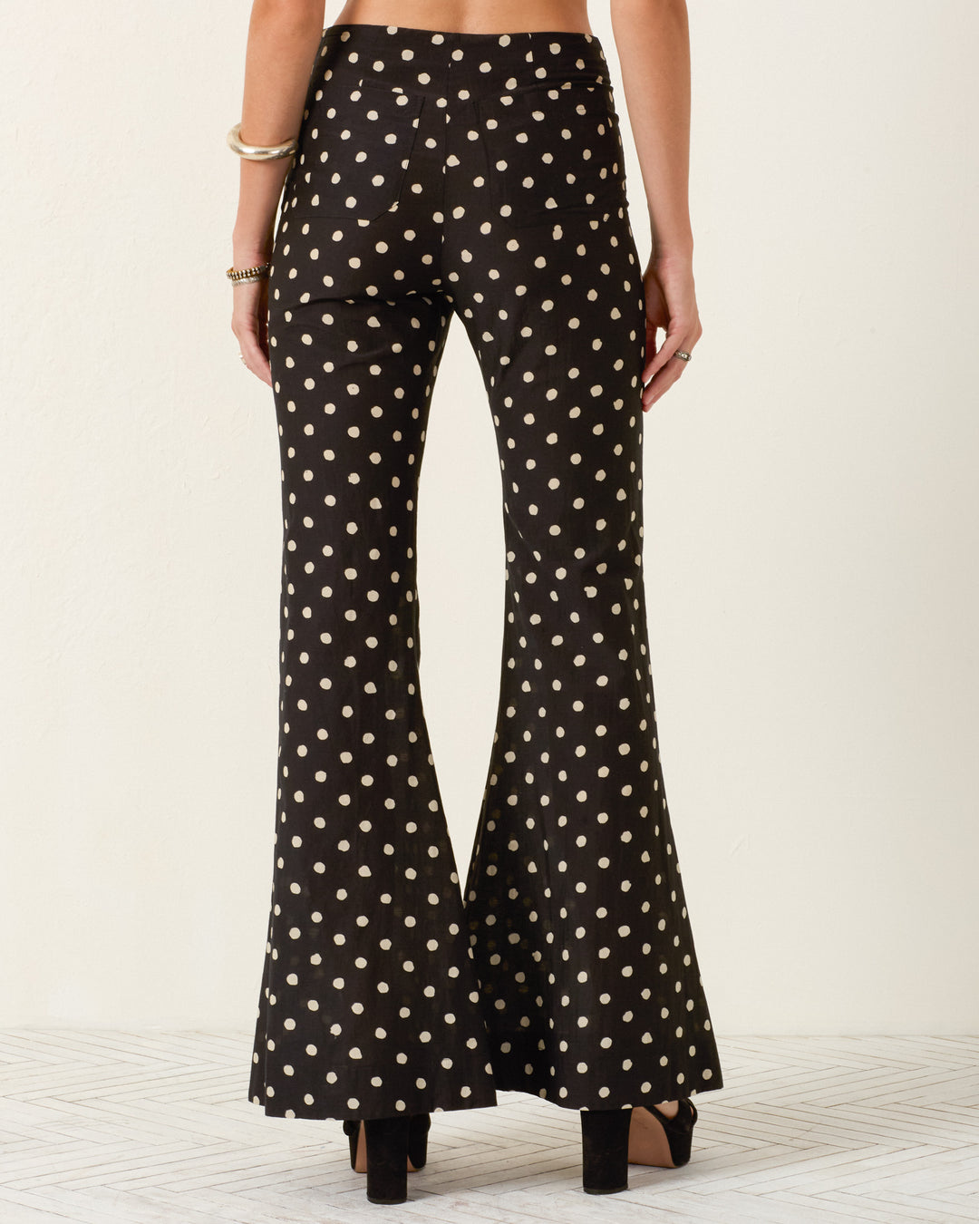A person is seen from the back wearing the Farah Blanc Speckle Pant, high-waisted with a black and white polka-dot flare design, styled with black platform heels and gold bracelets.