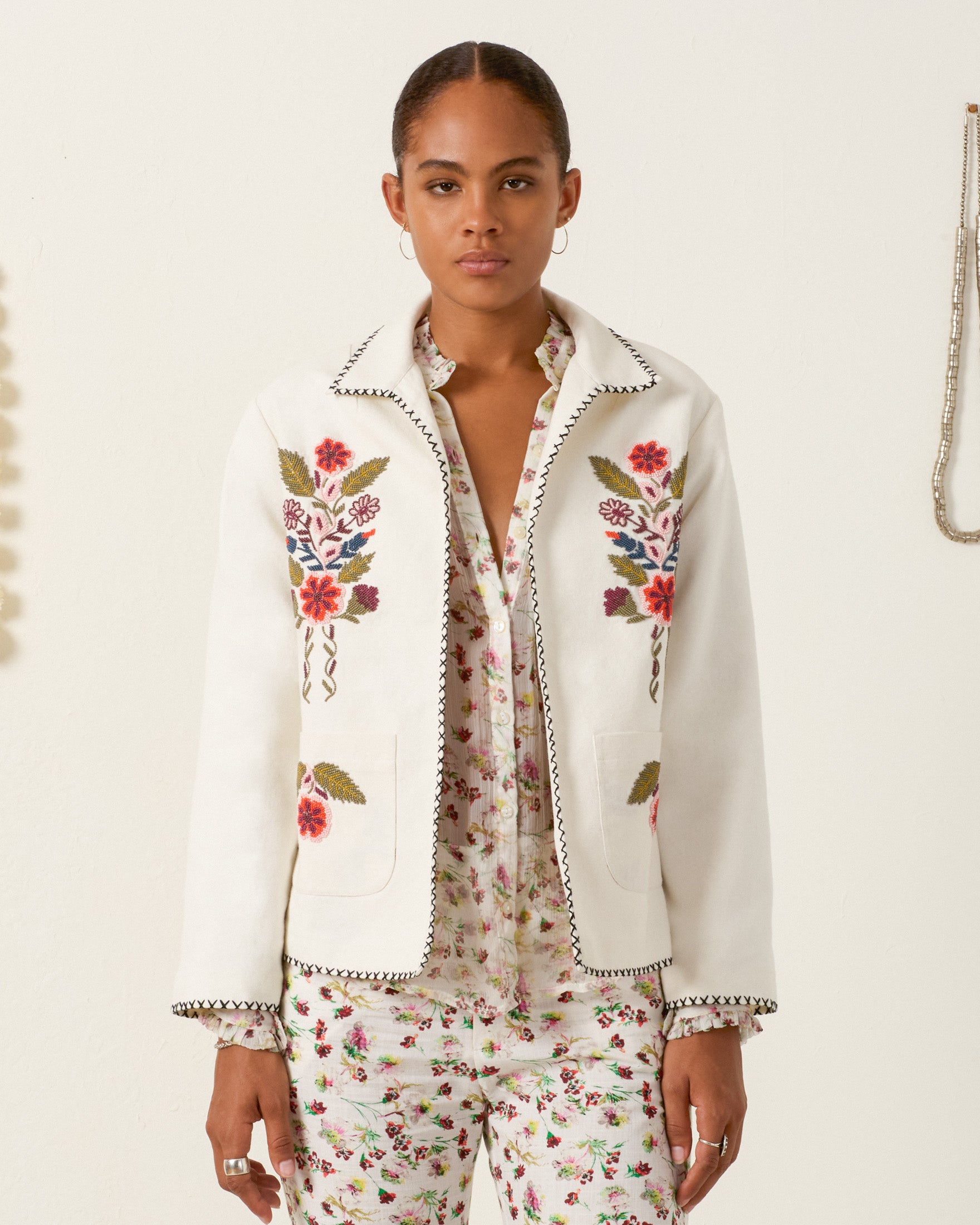A woman wears the Francis Heirloom Flora Jacket, featuring vintage-inspired colorful floral embroidery and beadwork, over a white textured cotton floral-patterned shirt and pants against a plain background.