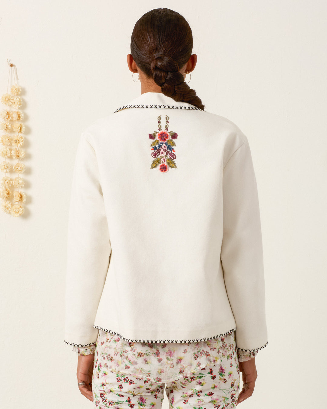 A person faces away, wearing the Francis Heirloom Flora Jacket with textured cotton, floral embroidery, and beadwork on the back, paired with floral-patterned pants. The plain background includes a small decorative item on the left wall.