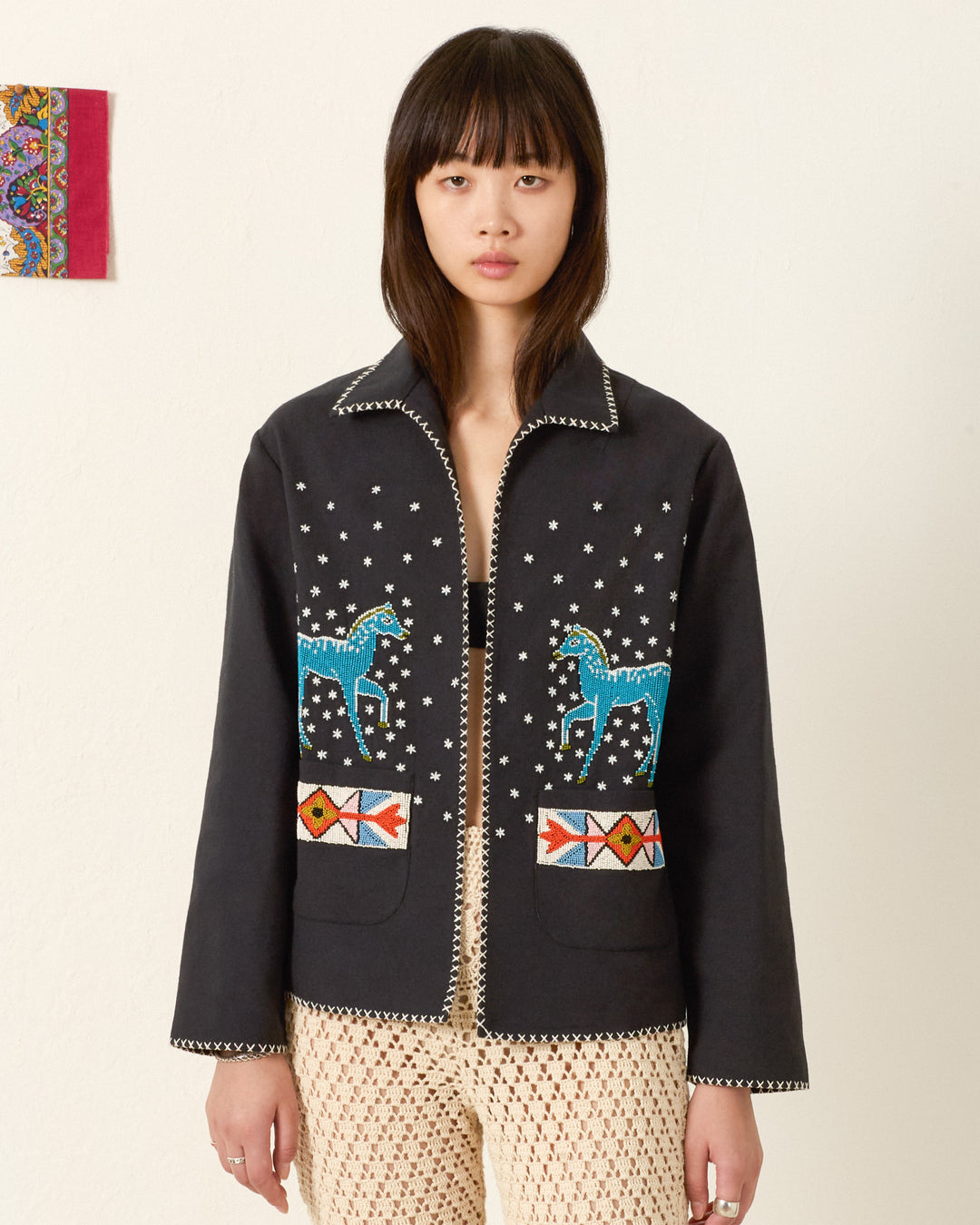 A woman stands against a plain wall wearing the Francis Fete Mare Jacket, featuring an embroidered horse motif, star patterns, and geometric designs over a cream crocheted outfit.