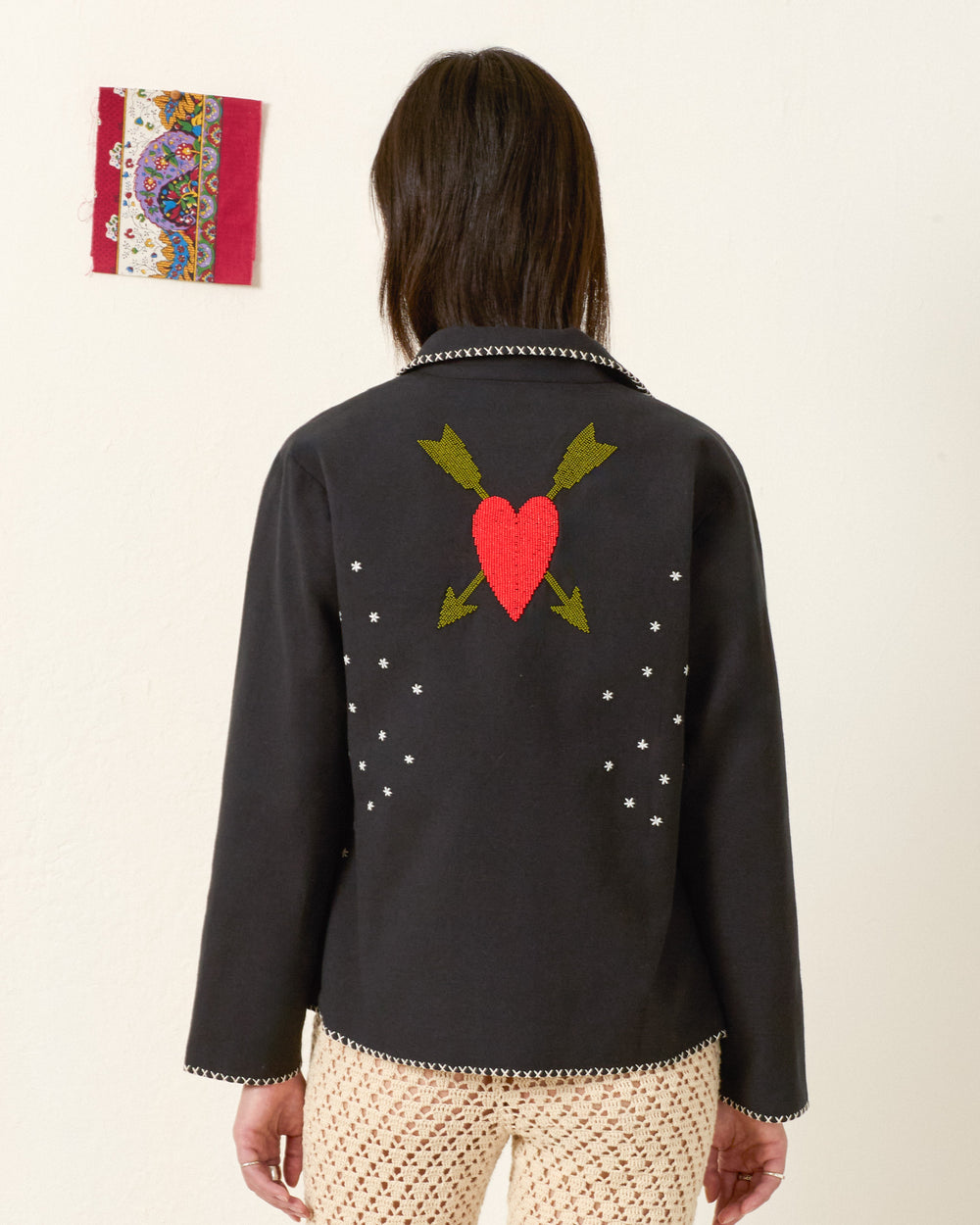 Person standing with back to camera, wearing the Francis Fete Mare Jacket featuring a red heart and arrow motif, white star details, and cream crochet pants. A small colorful textile artwork hangs on the wall.
