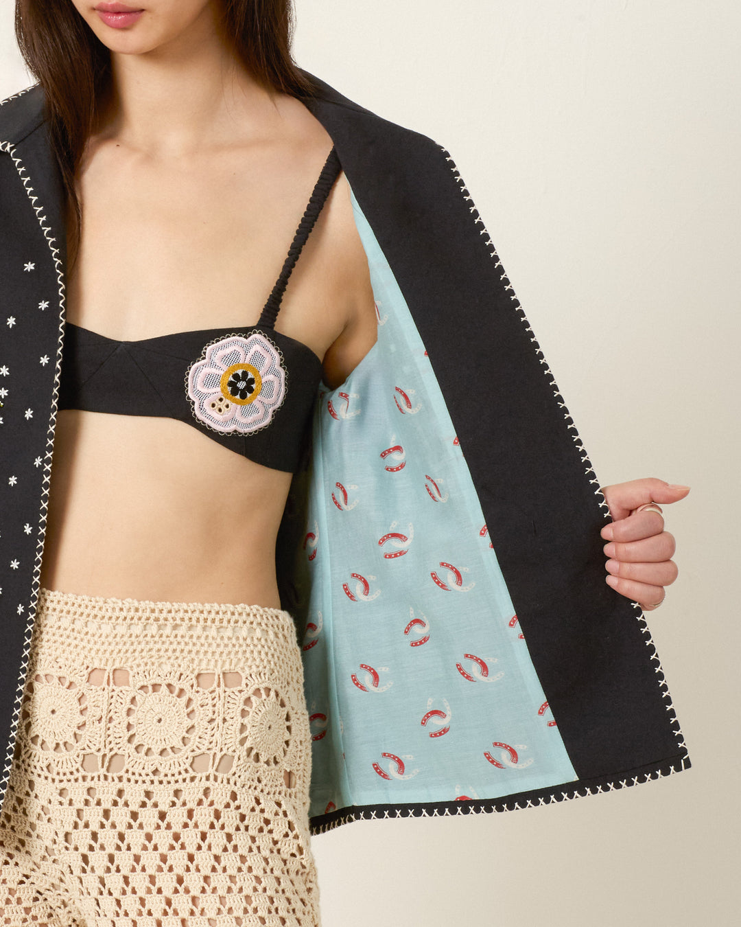 A woman wears a black bralette, beige crochet shorts, and the Francis Fete Mare Jacket, which features a light blue patterned lining. She holds the jacket open to reveal its intricate interior detail.