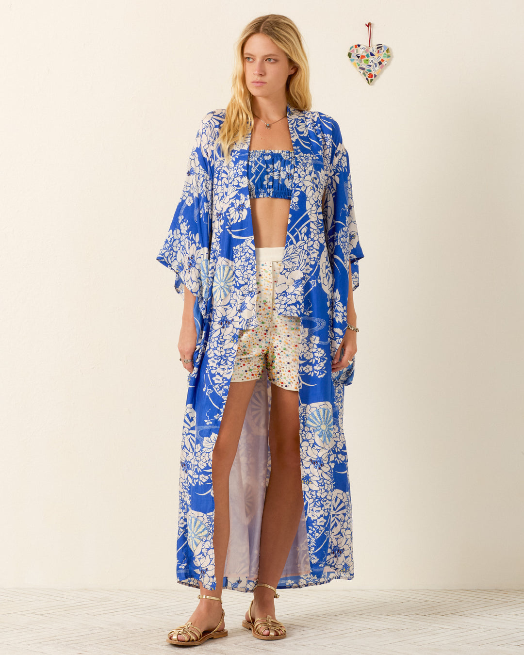 A woman stands indoors by a heart-shaped wall decor, wearing the Grace Royal Spoke Kimono Dress in a blue and white floral print, paired with cream printed shorts, a matching crop top, and sandals.