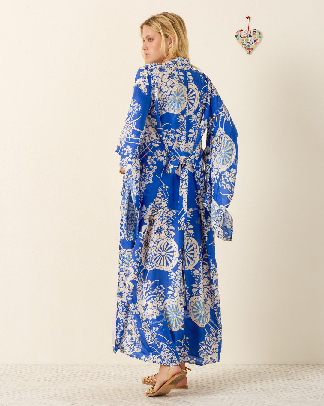 A person stands barefoot, facing away in a light room with a small heart on the wall, wearing the Grace Royal Spoke Kimono Dress—a long, hand-loomed silk wrap featuring a blue flower cart motif.