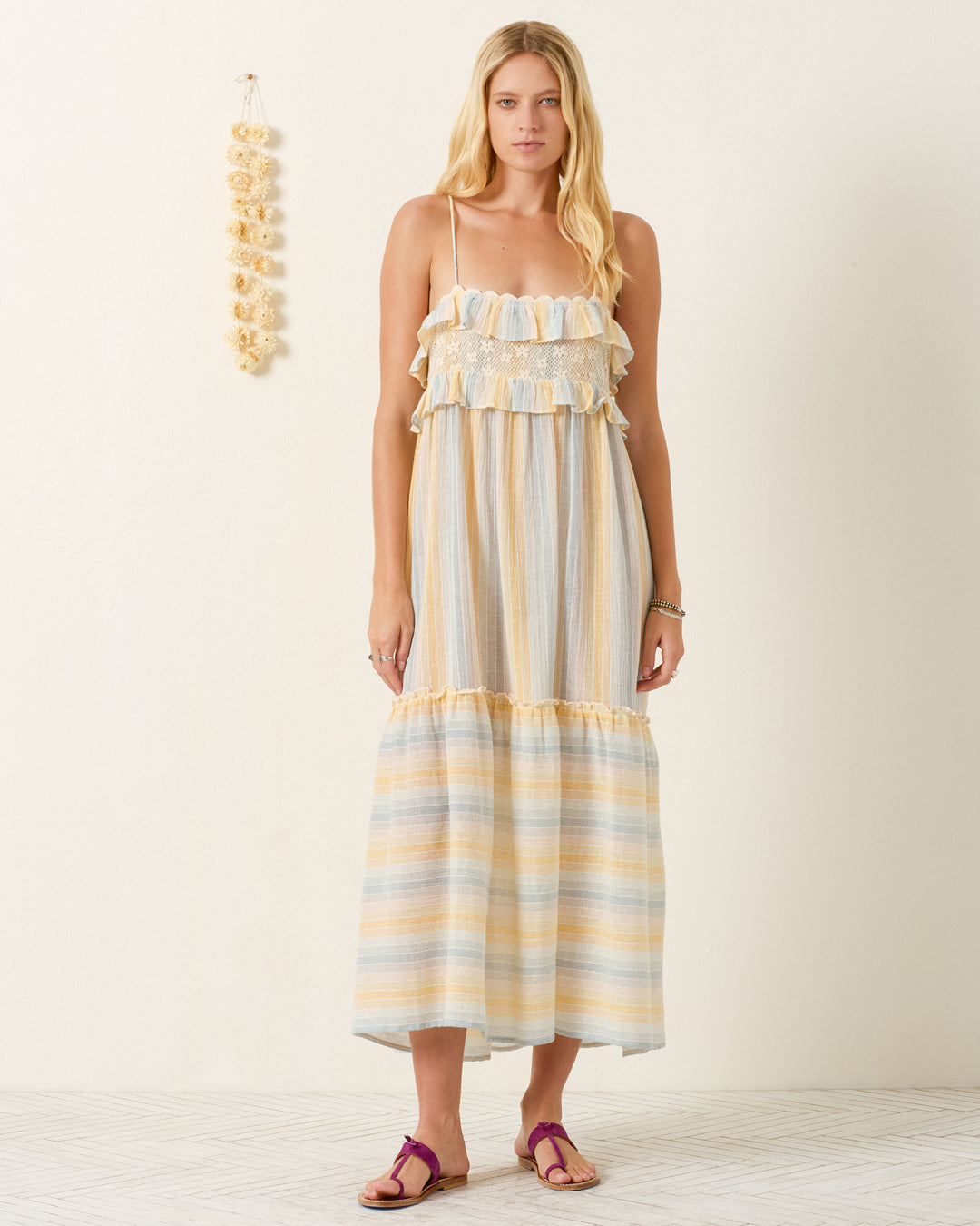 A woman stands indoors in the Ida Sunwash Stripe Dress, featuring gauzy cotton, ruffle details, and adjustable shoulder straps. She pairs it with purple sandals; a decorative hanging is visible on the wall behind her.