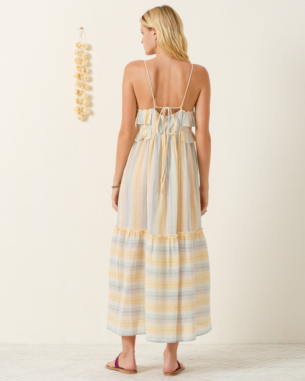 A woman with long blonde hair stands indoors, facing away from the camera, wearing the backless Ida Sunwash Stripe Dress with adjustable shoulder straps. The gauzy cotton fabric of her summer dress gives an airy feel to the scene.