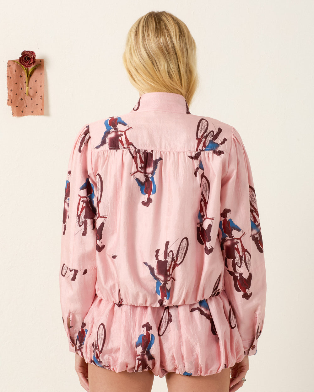 A blonde-haired person faces away, wearing the Jodie Parisian Pedal Jacket in pink with a vintage cyclist print. A small pink swatch with a rose and polka dots is pinned to the wall.