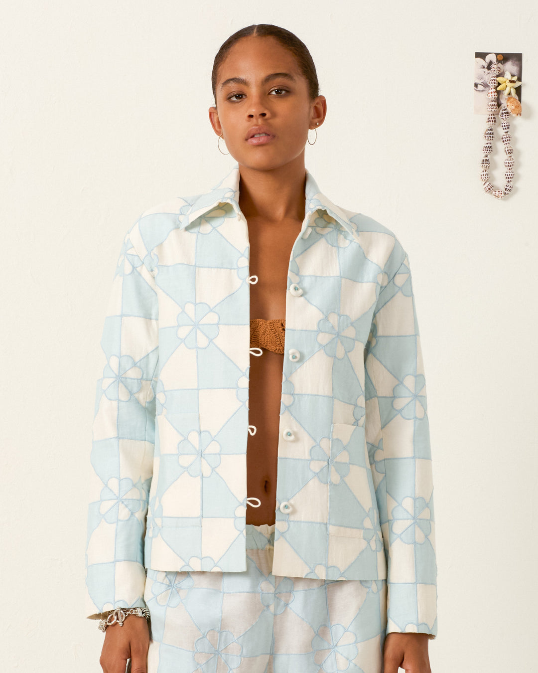 A woman stands against a plain wall, wearing the Jude Sky Jubilee Jacket—a cotton-silk blend in light blue and white checks with floral patterns—paired with a matching skirt.