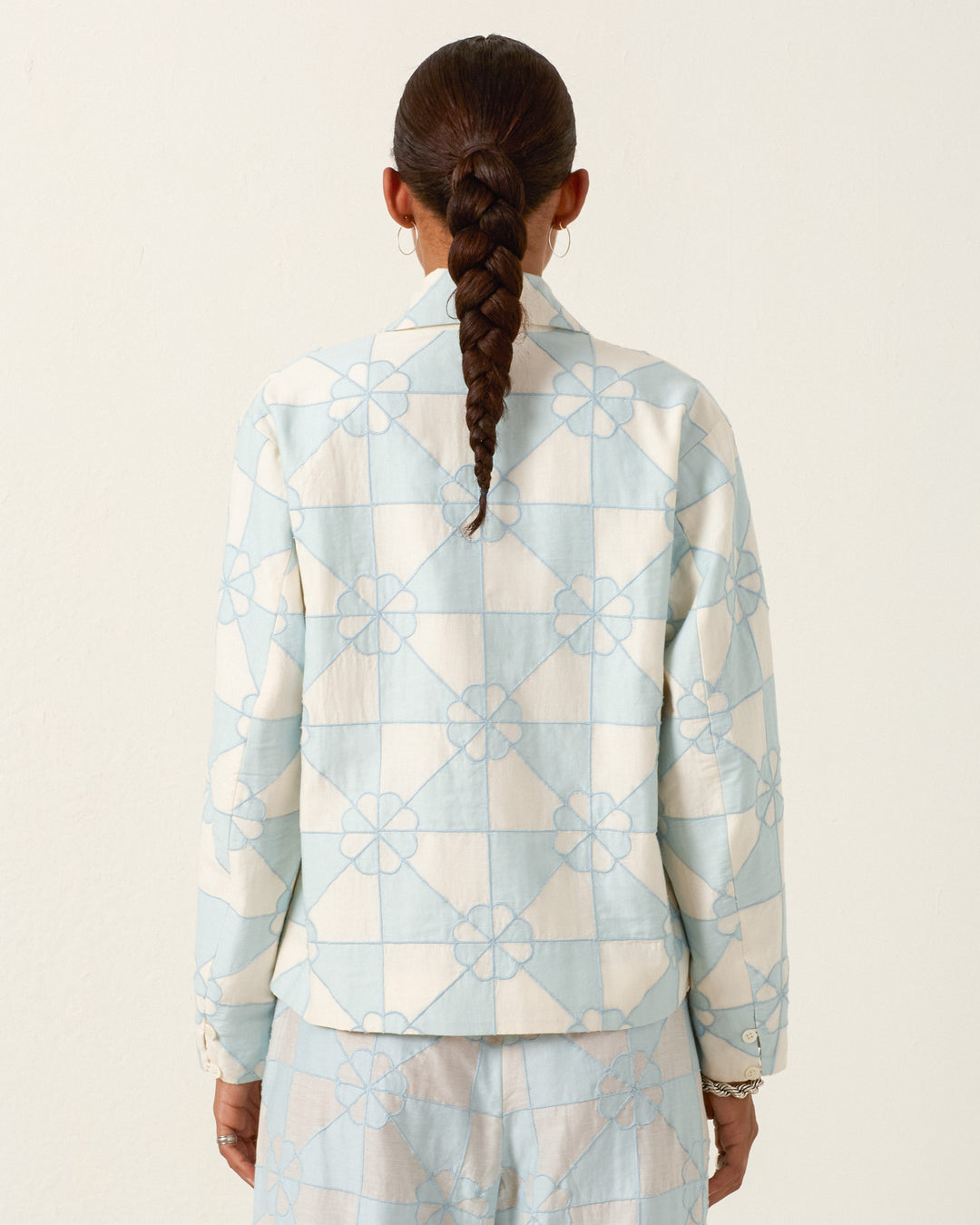 A person with a long braid, wearing the Jude Sky Jubilee Jacket—a light blue and white patterned cotton-silk blend—stands facing away against a plain background.