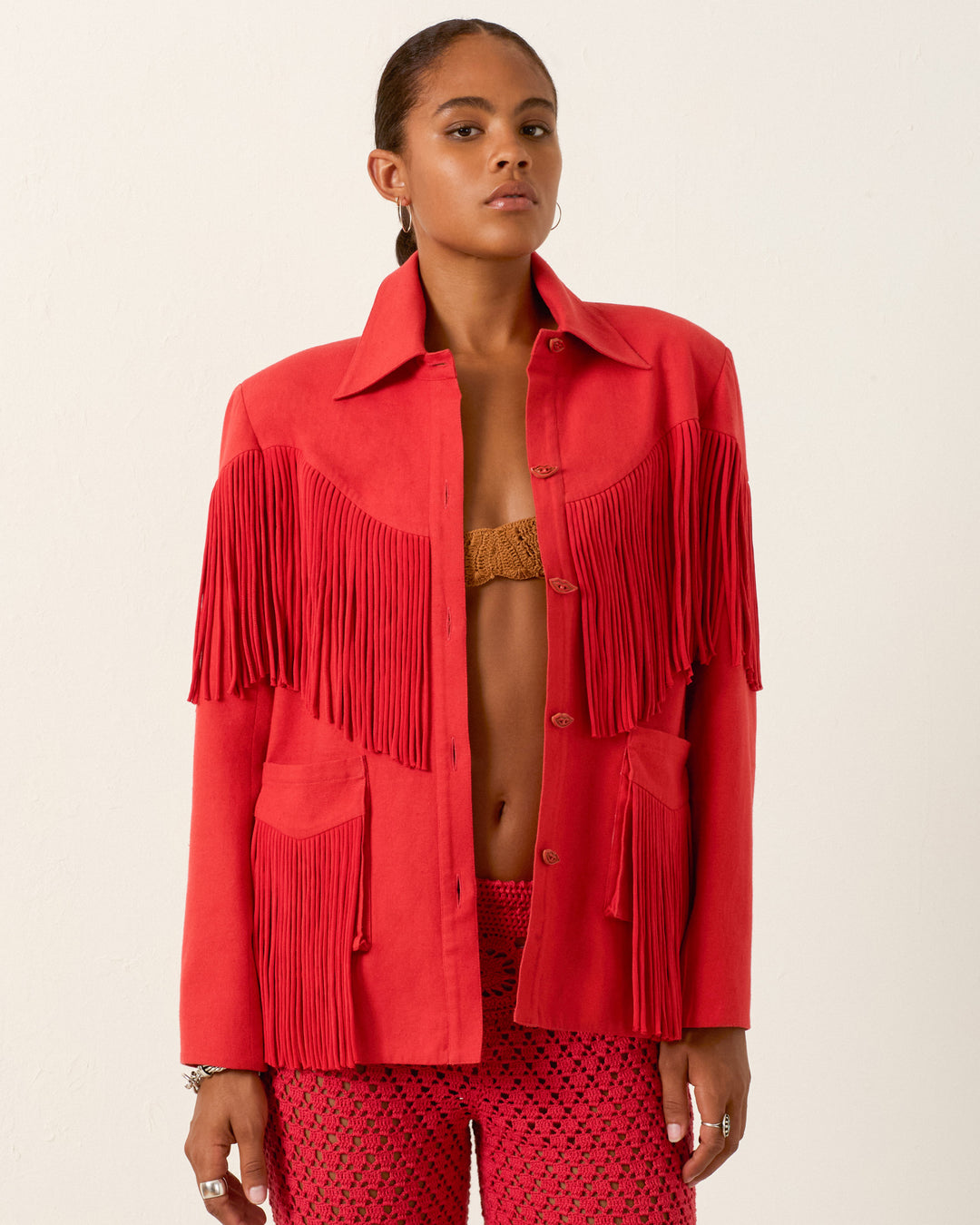 Wearing the handmade Jude Ibis Fringe Jacket, styled over a brown top and red patterned pants, a person stands against a plain light background.