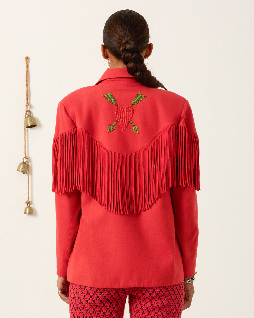 A person with a braided ponytail wears the Jude Ibis Fringe Jacket—red, Western-inspired, with an embroidered heart and arrows, plus handmade silk fringe—and patterned red pants. Three brass bells hang on the wall.