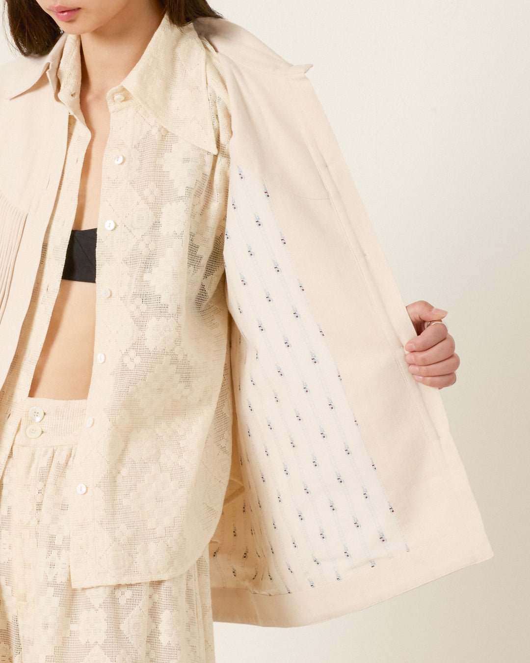 A person models a cream lace shirt and matching shorts with the Jude Dune Fringe Jacket in light beige draped over their shoulders, revealing its patterned lining.
