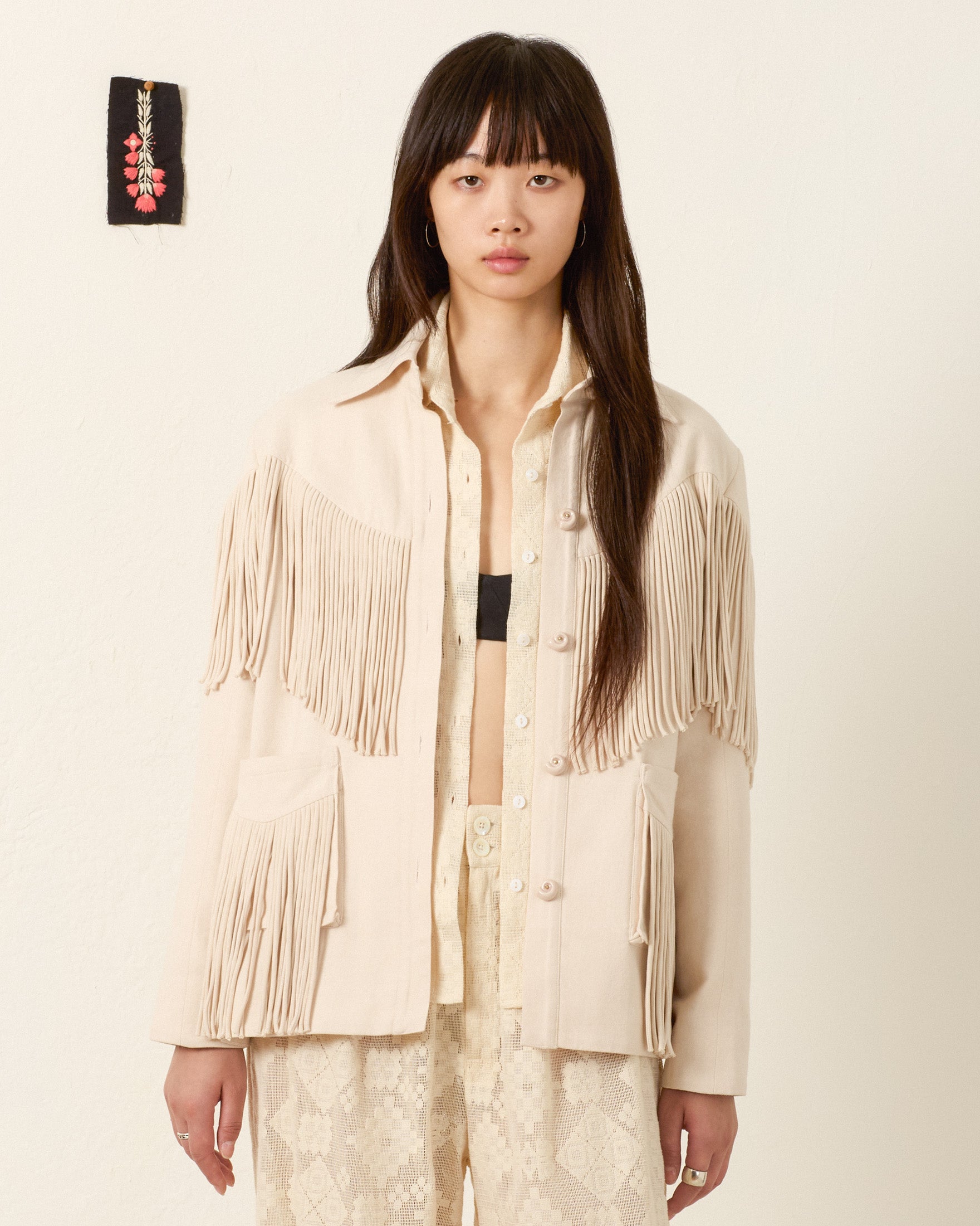 A woman poses against a plain wall, wearing the Jude Dune Fringe Jacket in cream silk over patterned pants and a partially unbuttoned shirt, with a small decorative piece on the wall behind her.