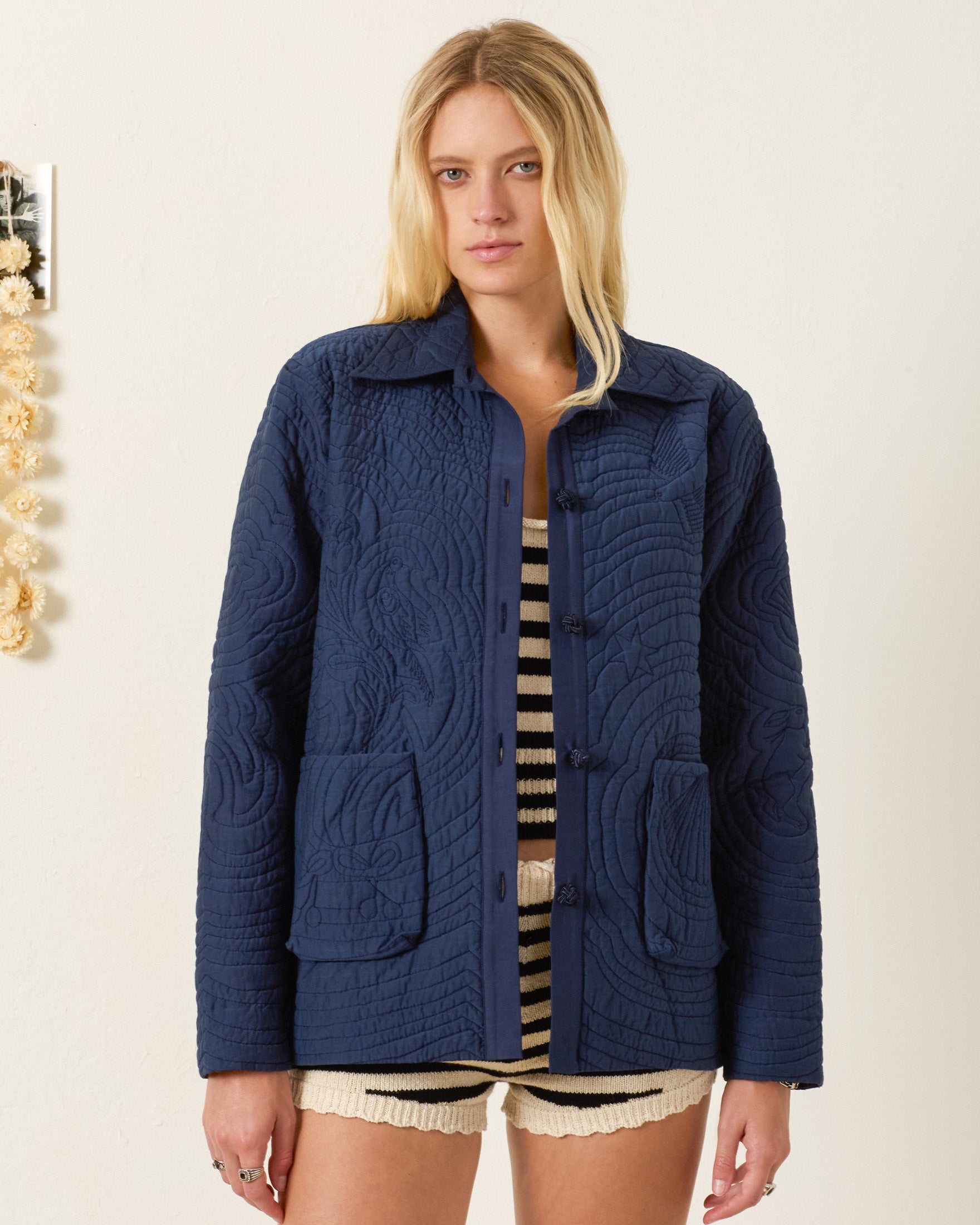 A woman with long blonde hair wears the Jude Midnight Reverie Jacket over a black and white striped top and cream shorts, standing against a plain background.