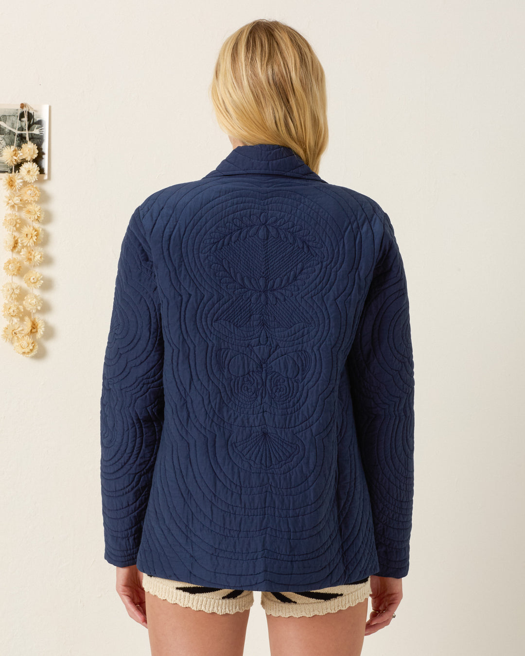 A person with long blonde hair wears the Jude Midnight Reverie Jacket, navy blue with quilted motifs, and cream shorts, standing back to the camera against a light wall.